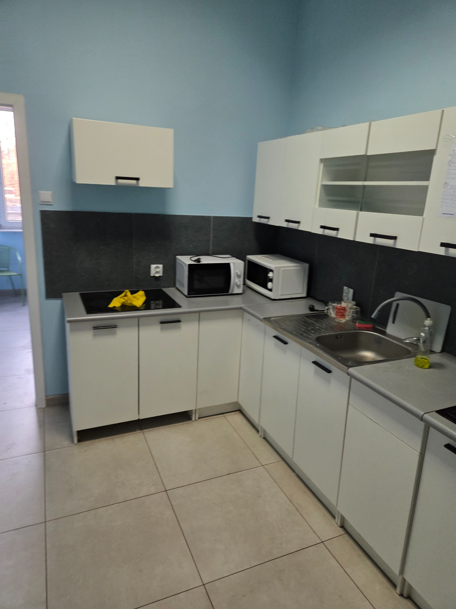 Jasna kuchnia w wynajmowanym mieszkaniu: białe szafki, szary blat, kuchenka indukcyjna z żółtą ścierką, mikrofalówka i zlewozmywak. Błękitne ściany i płytki podłogowe.