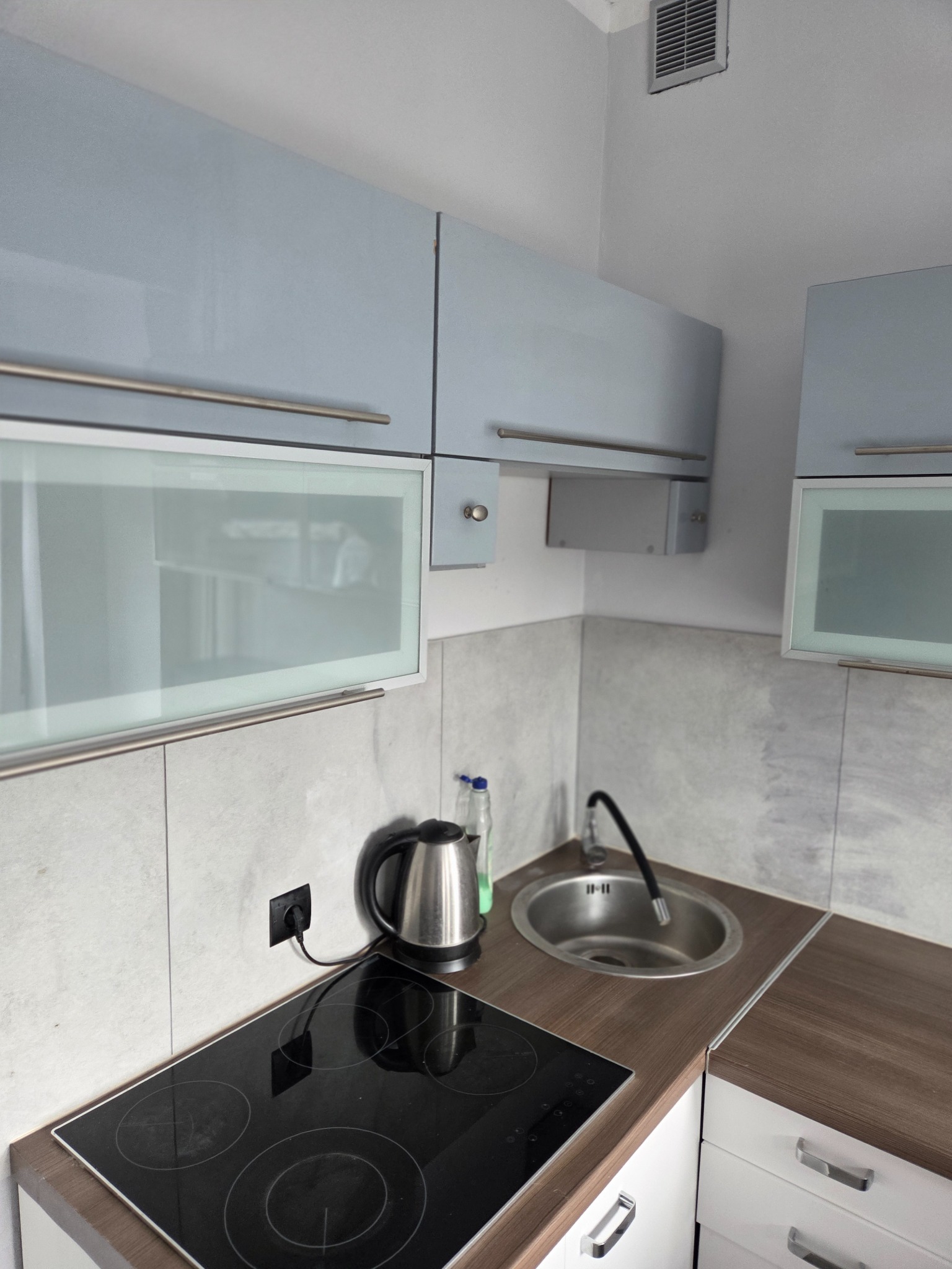 Kompaktowa kuchnia w wynajmowanym mieszkaniu: płyta indukcyjna, zlew, szafki z jasnoniebieskimi frontami i szklanymi wstawkami. Minimalistyczny design.