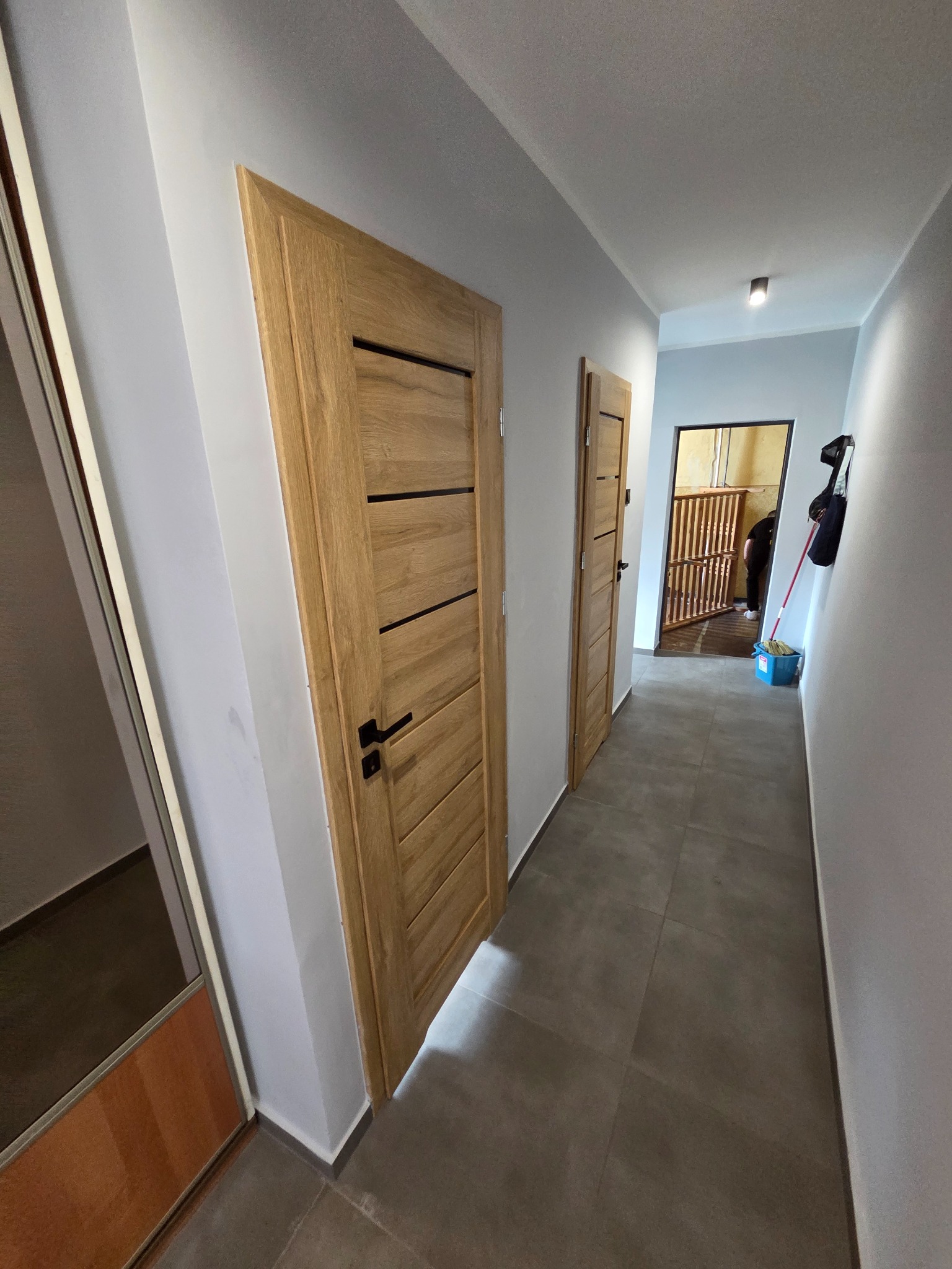 Korytarz z dwiema drewnianymi drzwiami z czarnymi klamkami, szarą podłogą i białymi ścianami. W tle widoczna osoba i wiadro z mopem.