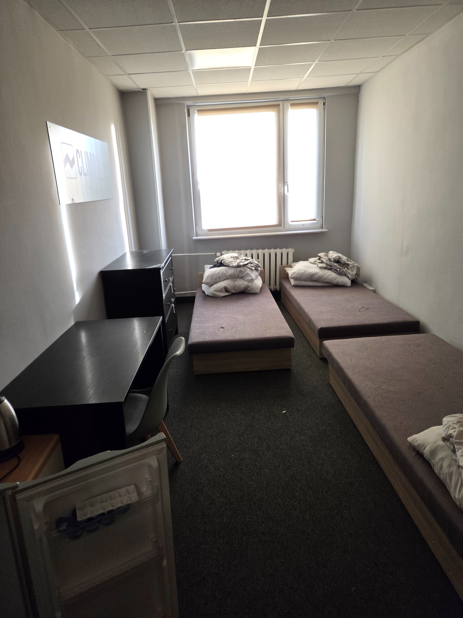 Pokój z trzema łóżkami, biurkiem, komodą i małą lodówką. Jasne światło wpada przez okno. Minimalistyczne wnętrze, potencjalnie pokój do wynajęcia.