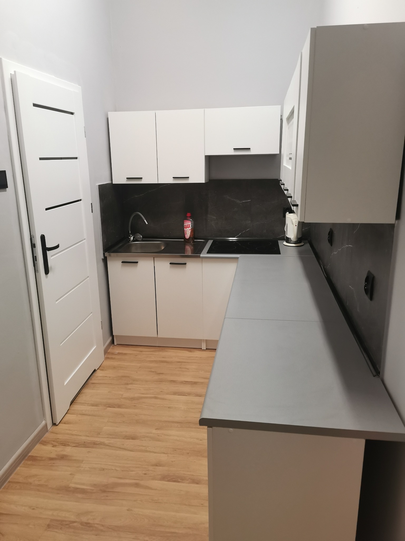 Nowoczesna, jasna kuchnia z białymi szafkami i szarym blatem. Widoczny zlew, płyta indukcyjna i drewniana podłoga. Minimalistyczny design, idealny do wynajmowanego mieszkania.