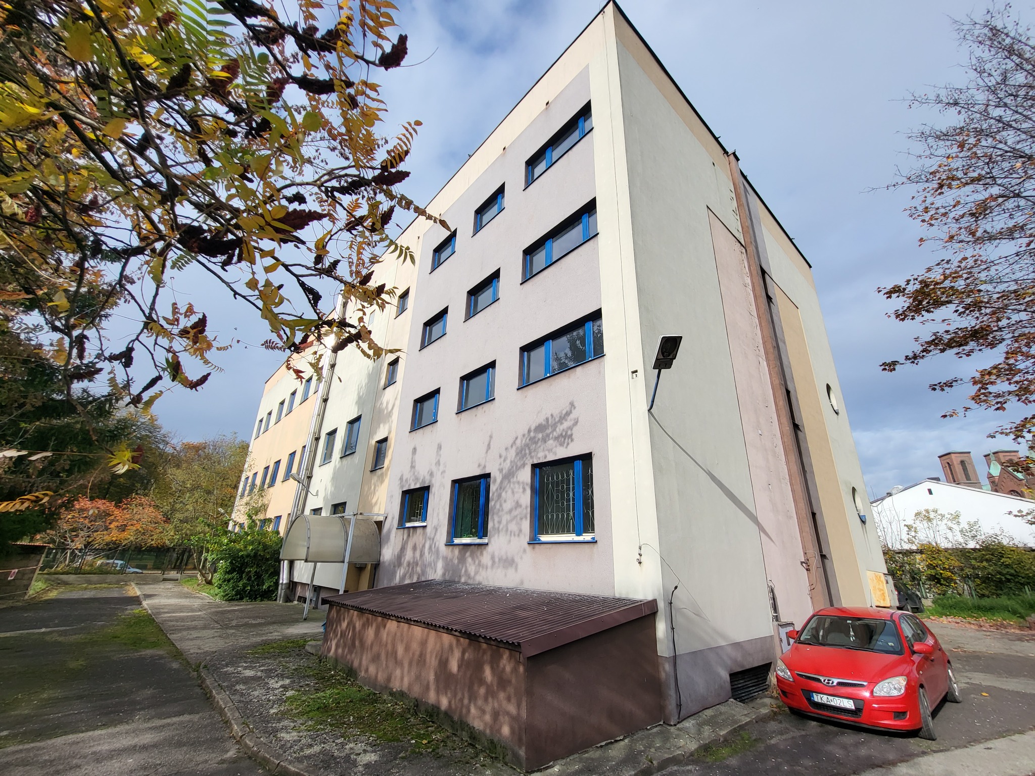 Wielopiętrowy budynek mieszkalny z niebieskimi ramami okiennymi, widziany z dołu, z czerwonym samochodem zaparkowanym obok. Jesienne drzewa w tle.