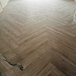 Aleks Batiment Aleksander Pruś - Ułożona podłoga z paneli w jodełkę, widoczne niedokończone gniazdko elektryczne z wystającymi kablami, tło stanowią okno i fragment ściany.