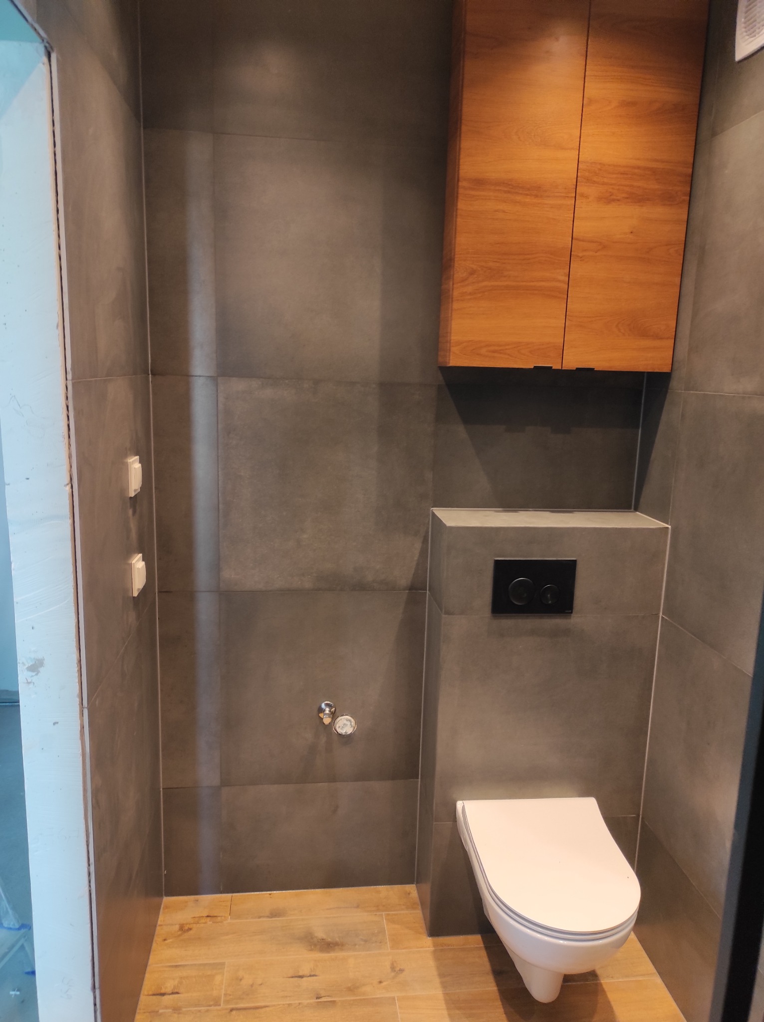 Nowoczesna łazienka z szarymi płytkami wielkoformatowymi, podwieszaną toaletą z czarnym przyciskiem, drewnianą szafką wiszącą i podłogą imitującą drewno.