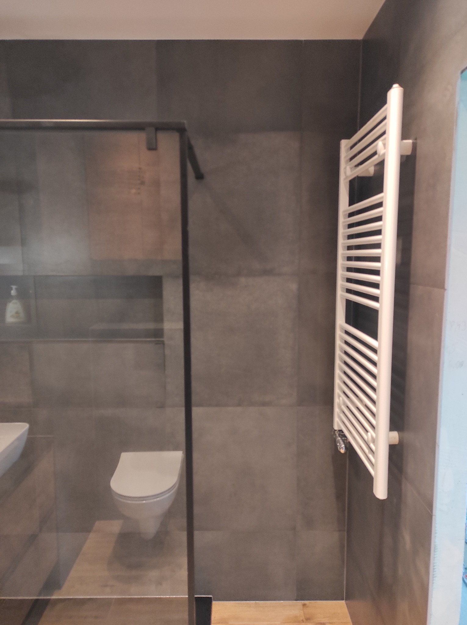 Nowoczesna łazienka z szarymi płytkami, kabiną prysznicową ze szkła, wiszącą toaletą i białym grzejnikiem drabinkowym.