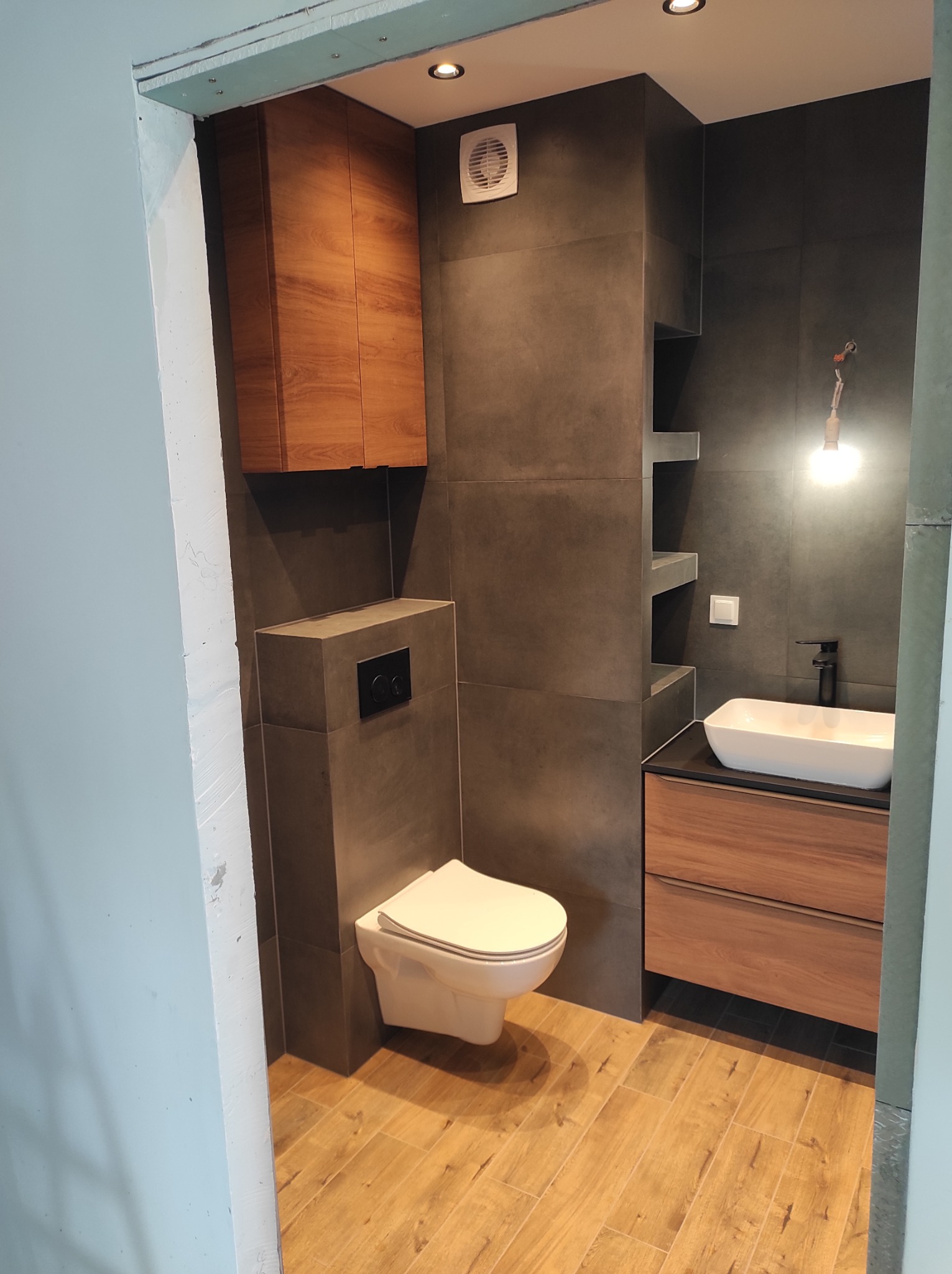 Nowoczesna łazienka z drewnianymi akcentami: szafka nad toaletą, umywalka nablatowa z szafką, szare płytki ścienne i podłoga imitująca drewno, widziane zza framugi drzwi.