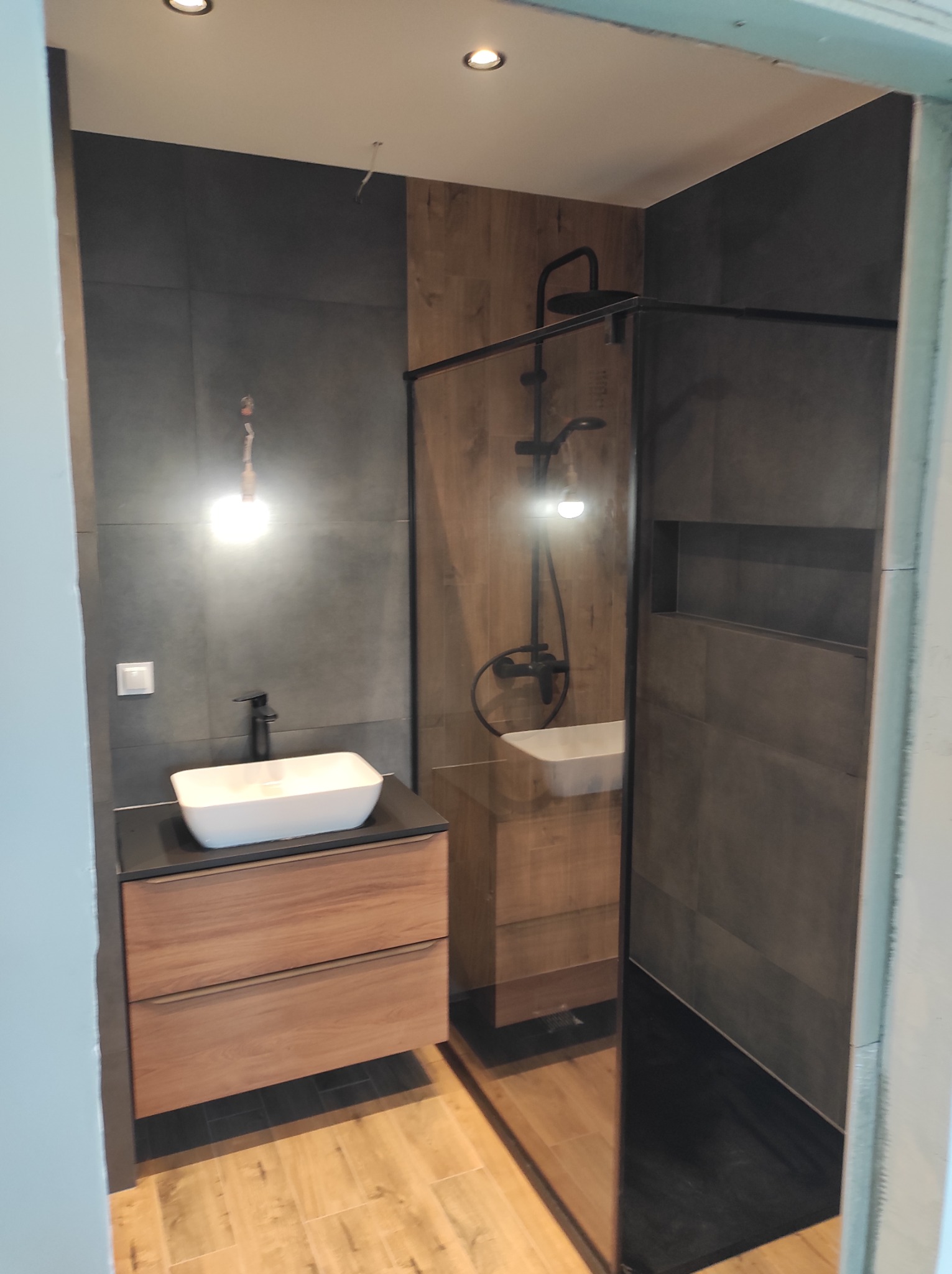 Nowoczesna łazienka z kabiną prysznicową typu walk-in, drewnopodobnymi i szarymi płytkami, umywalką nablatową i drewnianą szafką pod umywalką, oświetlona punktowo.