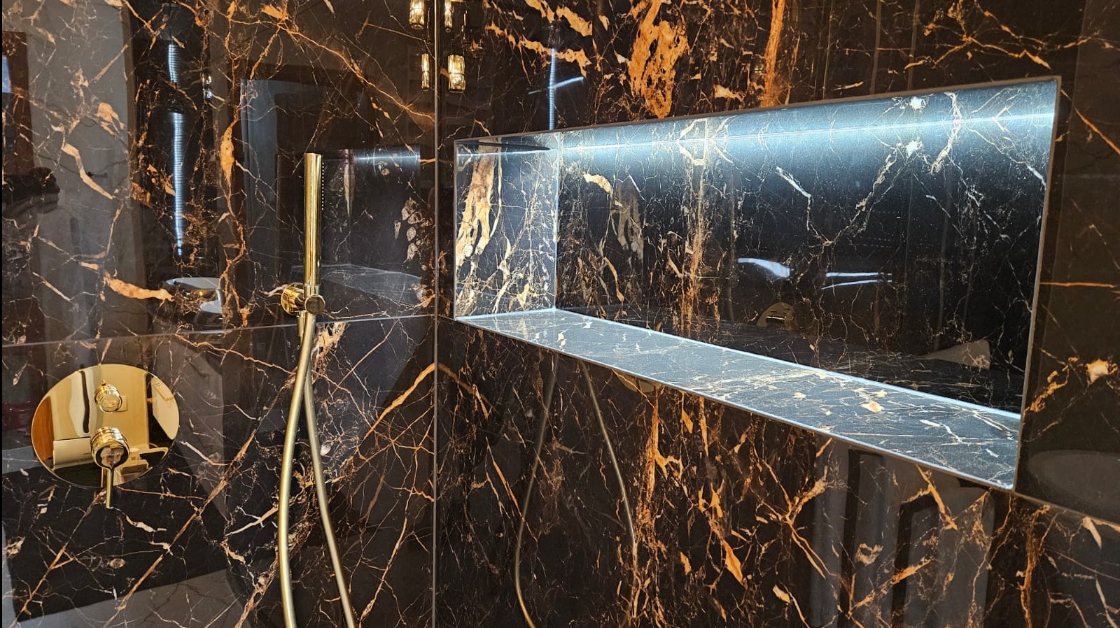 Luksusowa kabina prysznicowa w Krzeszowie wyłożona czarnym marmurem ze złotymi żyłami, z wnęką podświetloną ledami i złotą armaturą.
