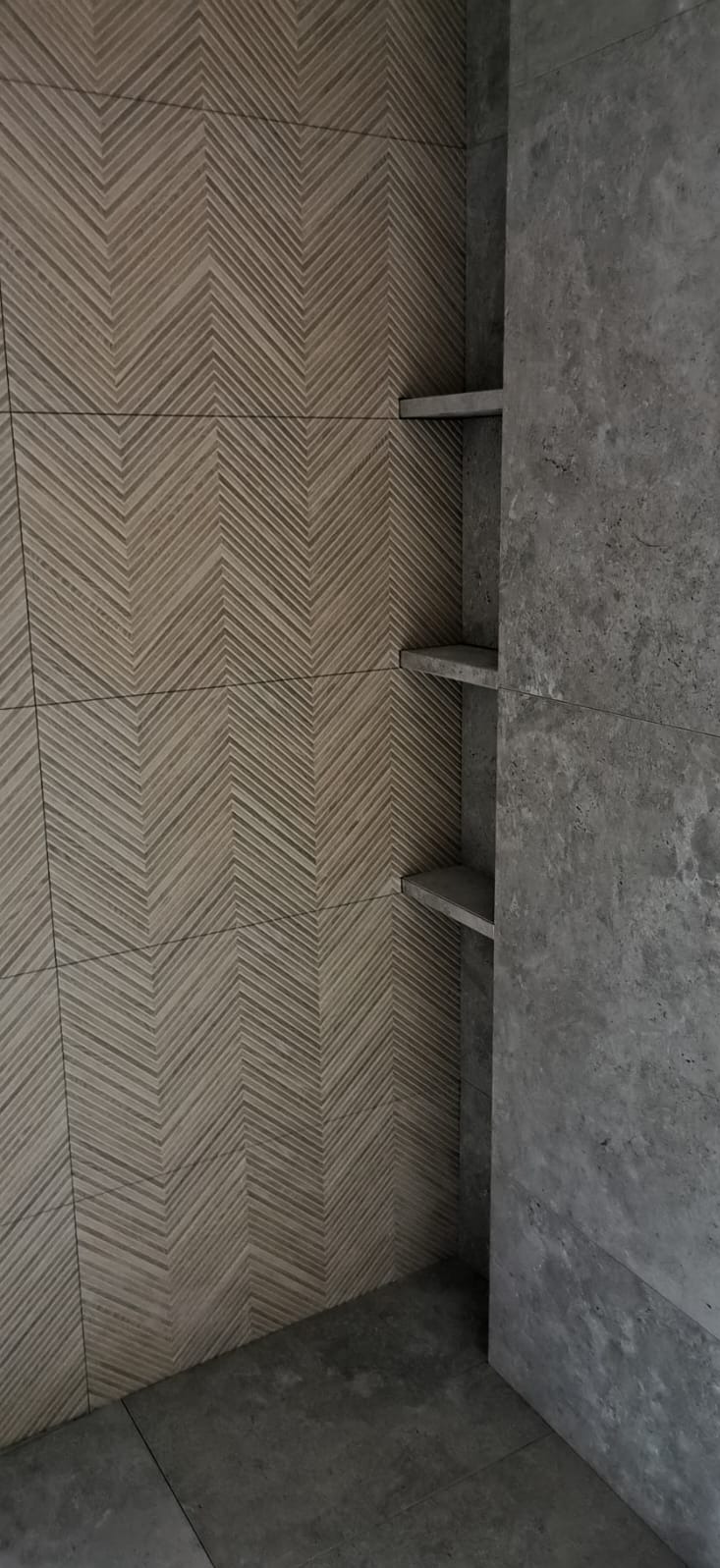 Wykończona ściana łazienki z płytek w jodełkę w odcieniach beżu, połączona z szarą, betonową ścianą z trzema wbudowanymi półkami.