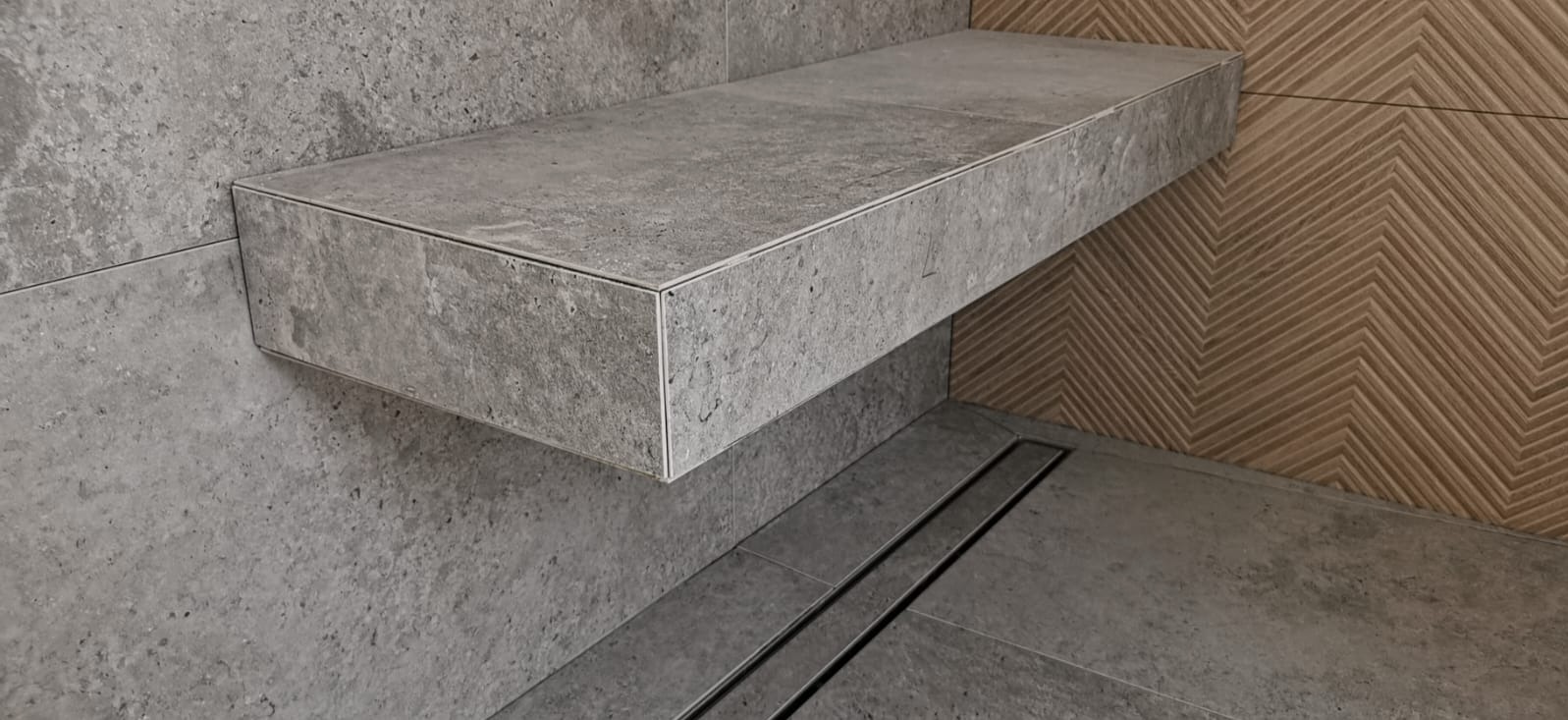 Półka z szarej płytki w nowoczesnej łazience z odpływem liniowym i dekoracyjną ścianą z wzorem jodełki.