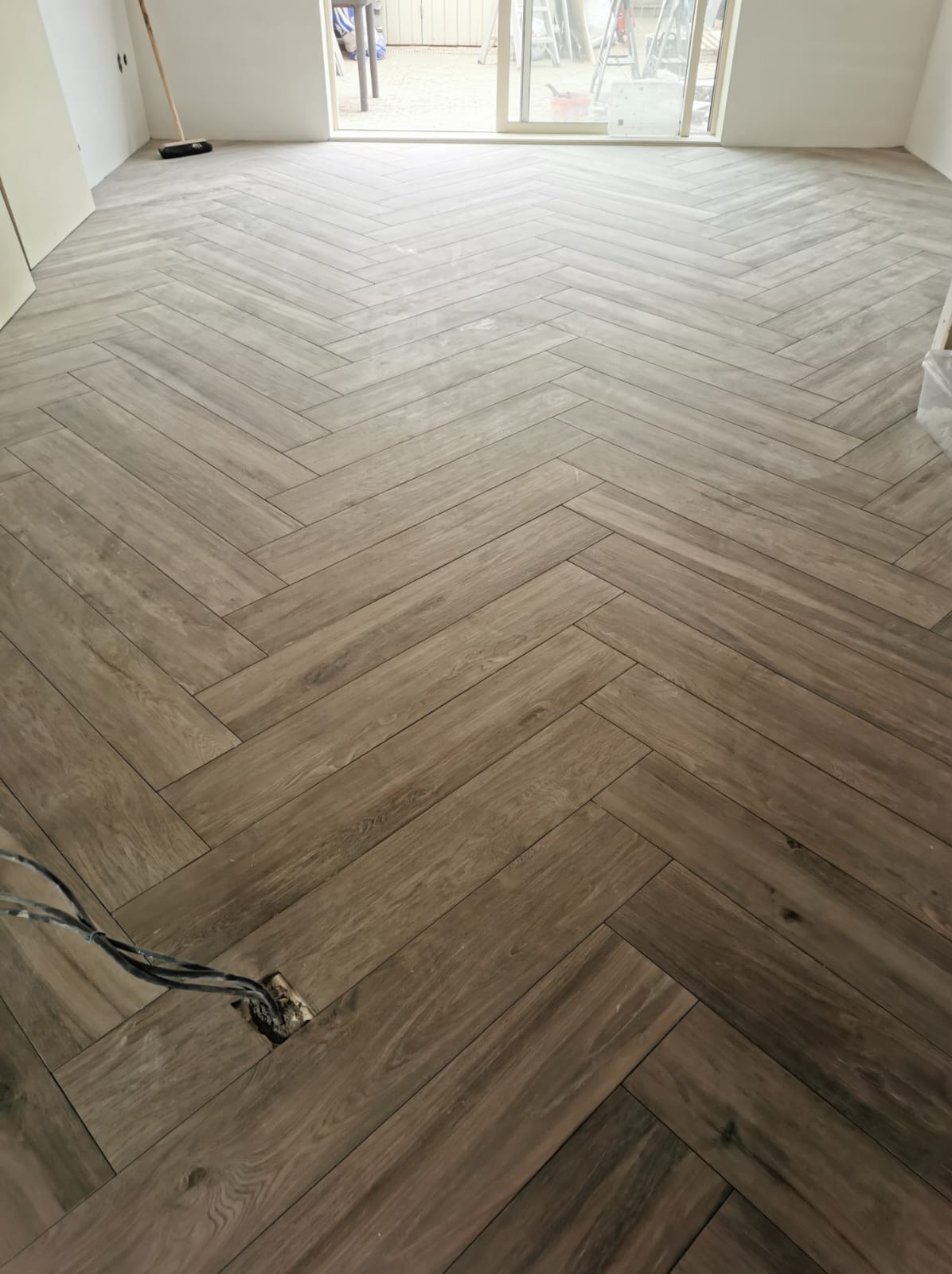 Ułożona podłoga z paneli w jodełkę, widoczne niedokończone gniazdko elektryczne z wystającymi kablami, tło stanowią okno i fragment ściany.