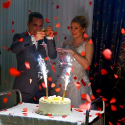 Uśmiechnięta para młoda wznosi toast za tort weselny ze sztucznymi ogniami, otoczona opadającymi konfetti w kształcie serc.