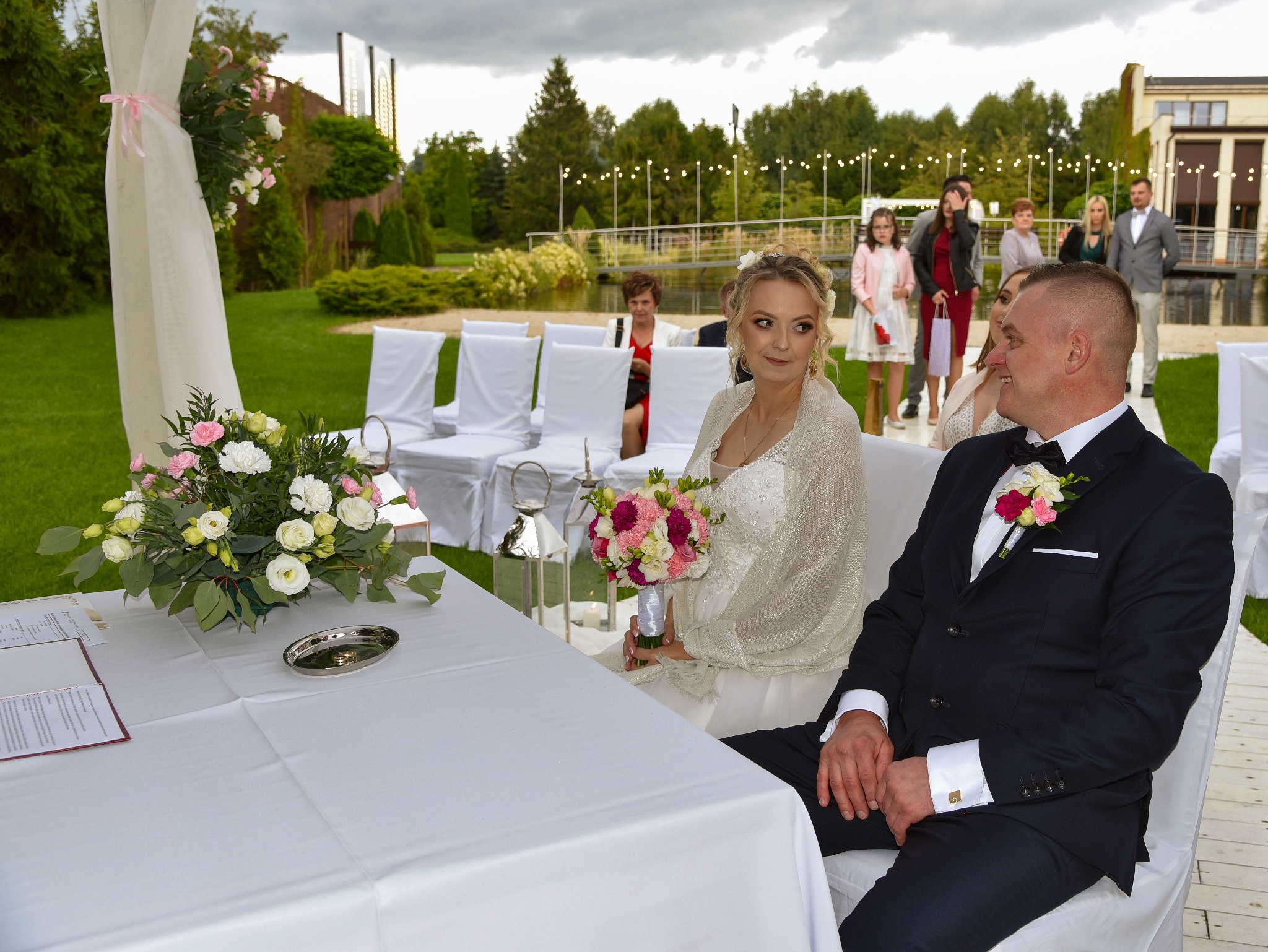 Para młoda siedzi przy stole podczas ceremonii ślubnej na zewnątrz, w tle goście i dekoracje.