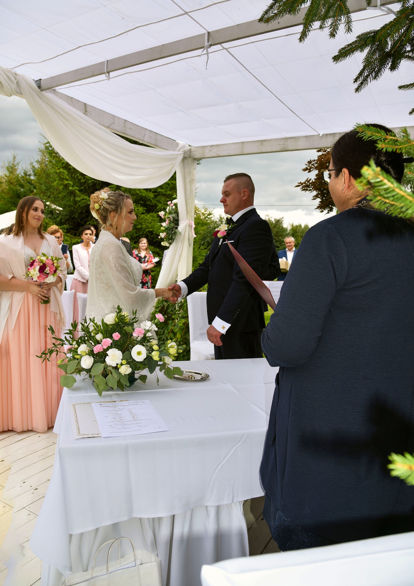 Para młoda wymienia uścisk dłoni podczas ceremonii ślubnej pod białym baldachimem, z bukietem kwiatów na stole i świadkami w tle.