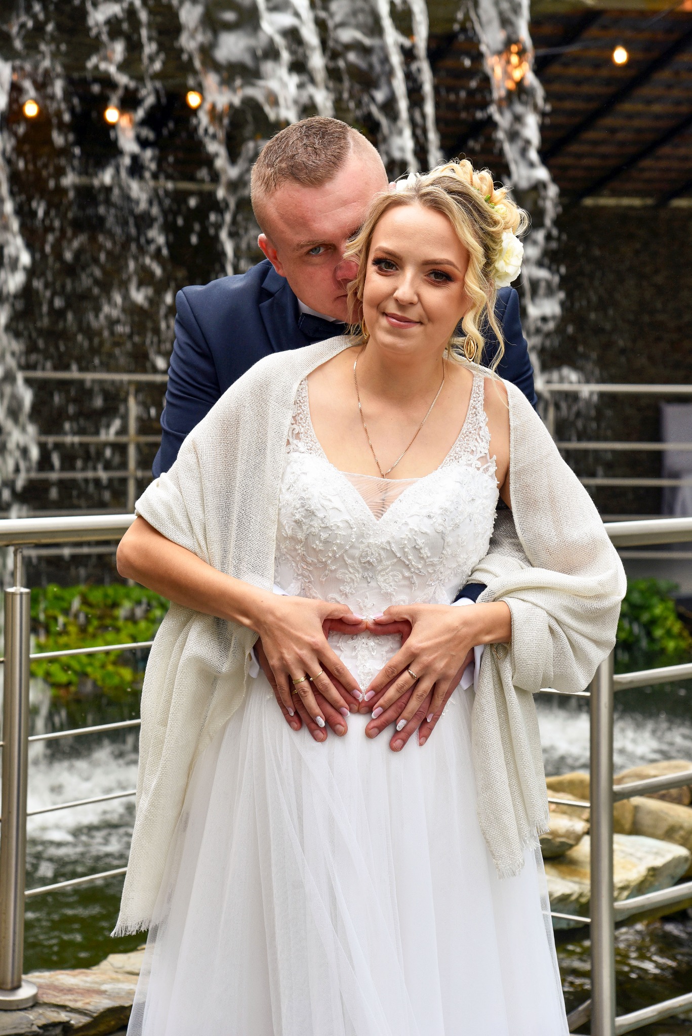 Para młoda w białej sukni i granatowym garniturze pozuje na tle wodospadu, dłońmi tworząc serce na brzuchu panny młodej.