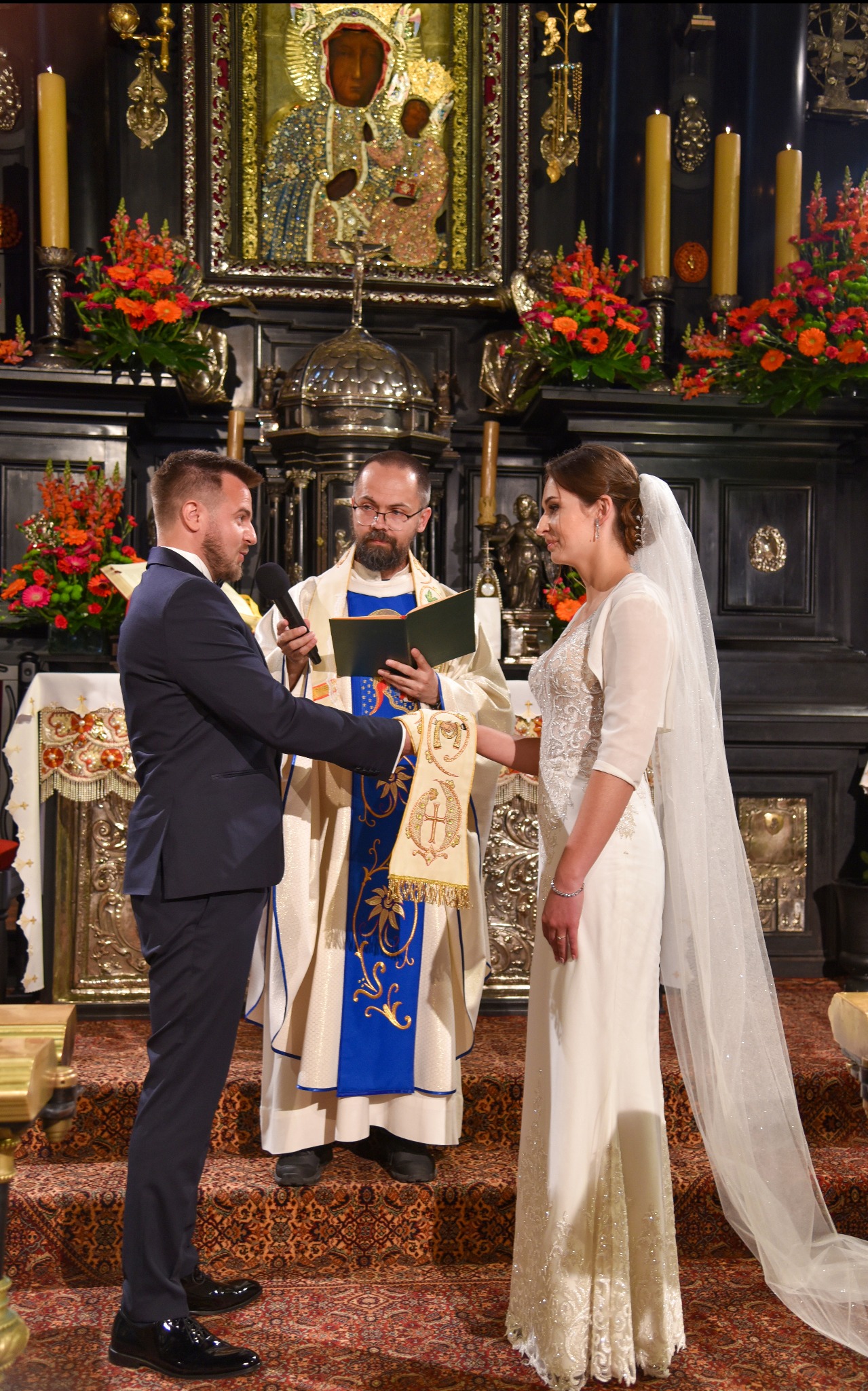 Para młoda podczas ceremonii ślubnej w kościele, ksiądz w ornacie z mikrofonem, bogato zdobiony ołtarz w tle.