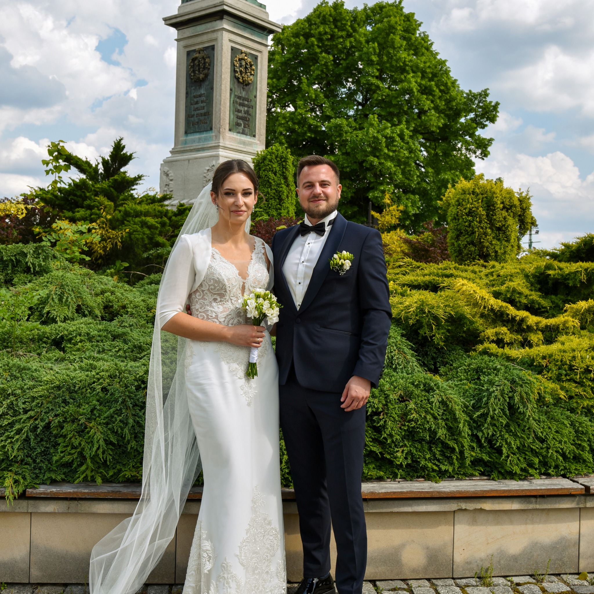 Para młoda w strojach ślubnych pozuje na tle pomnika i zieleni w plenerze.