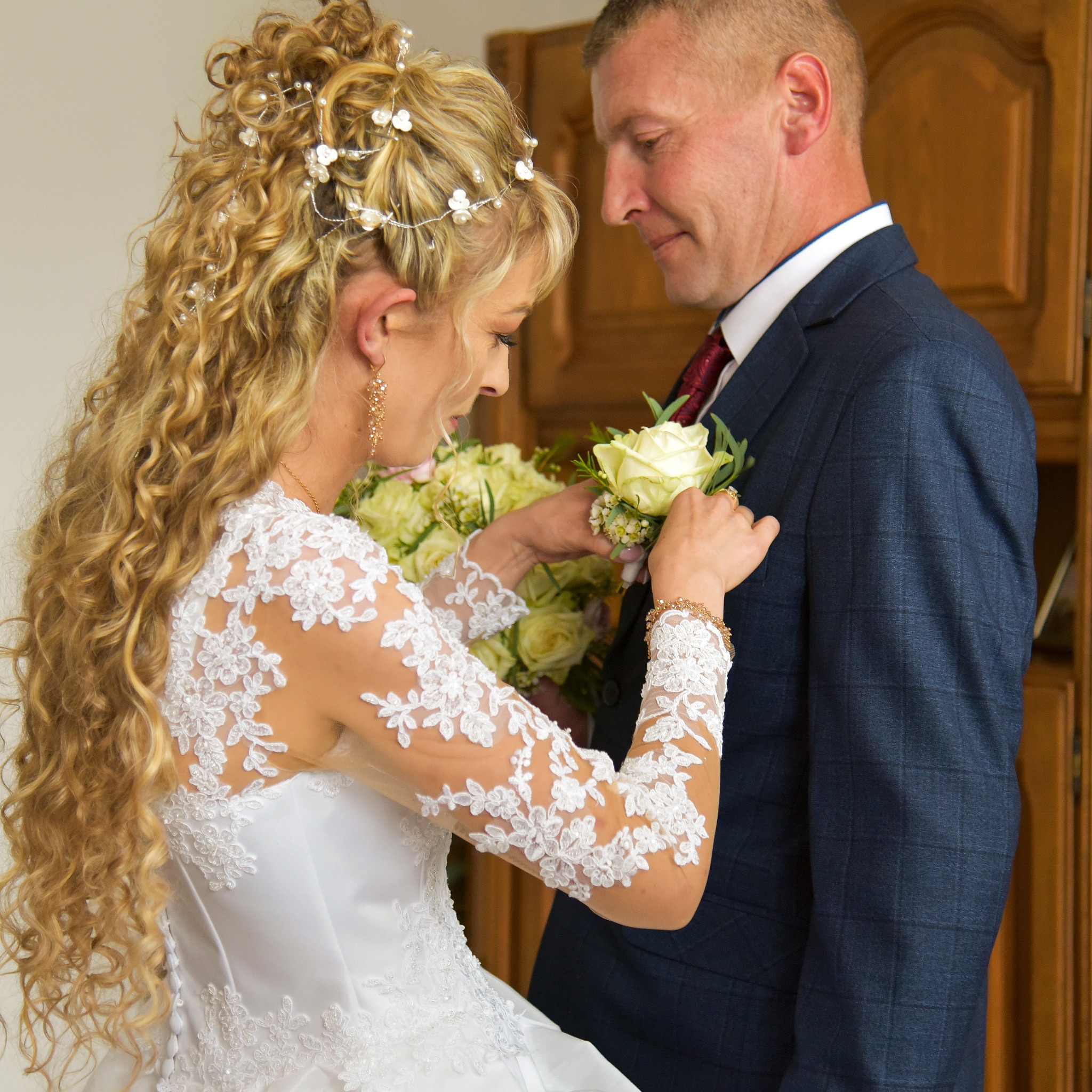 Panna młoda w koronkowej sukni przypina butonierkę z białej róży do klapy granatowego garnituru pana młodego.