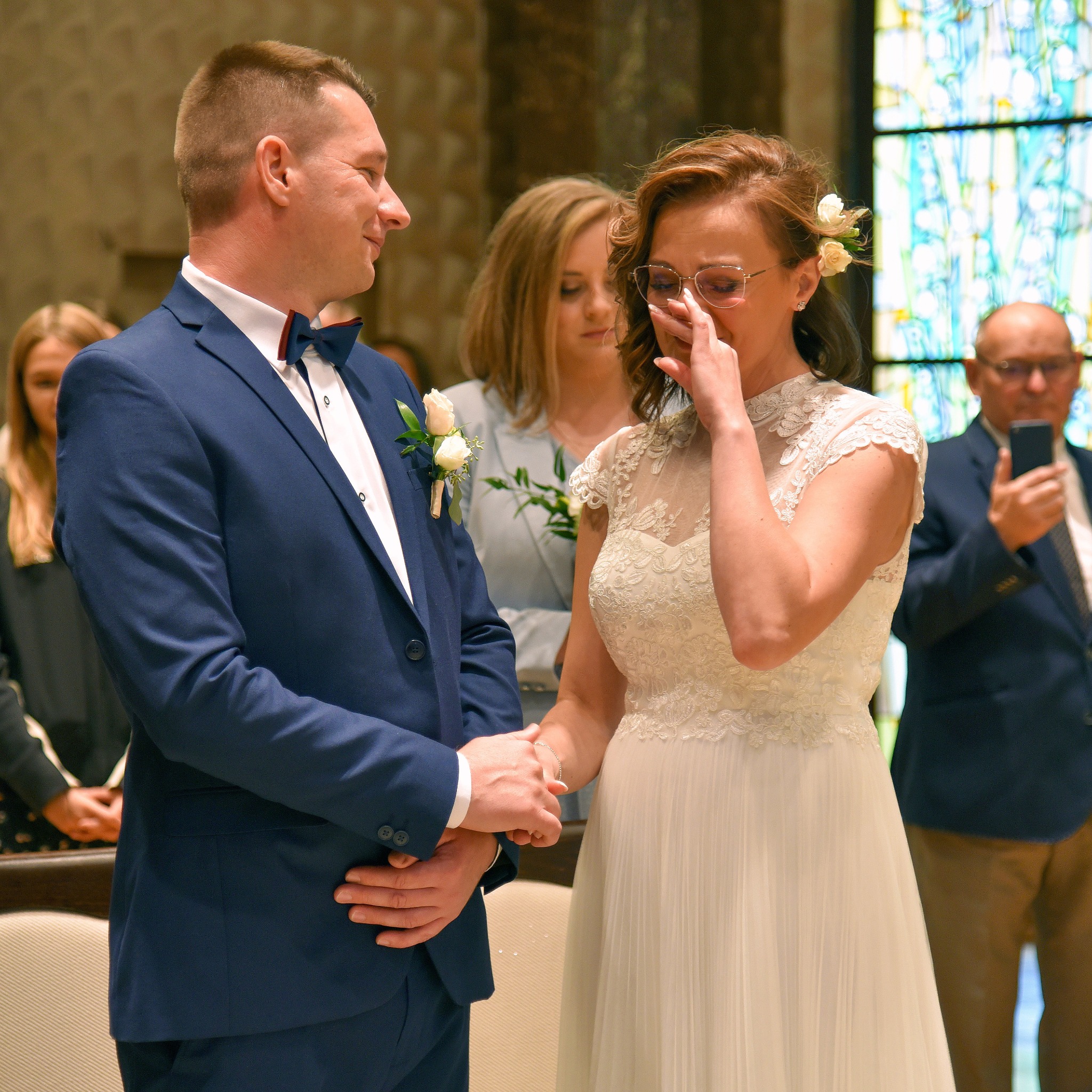 Panna młoda ociera łzy podczas ceremonii ślubnej, trzymając za rękę pana młodego w niebieskim garniturze. W tle witrażowe okno i goście.