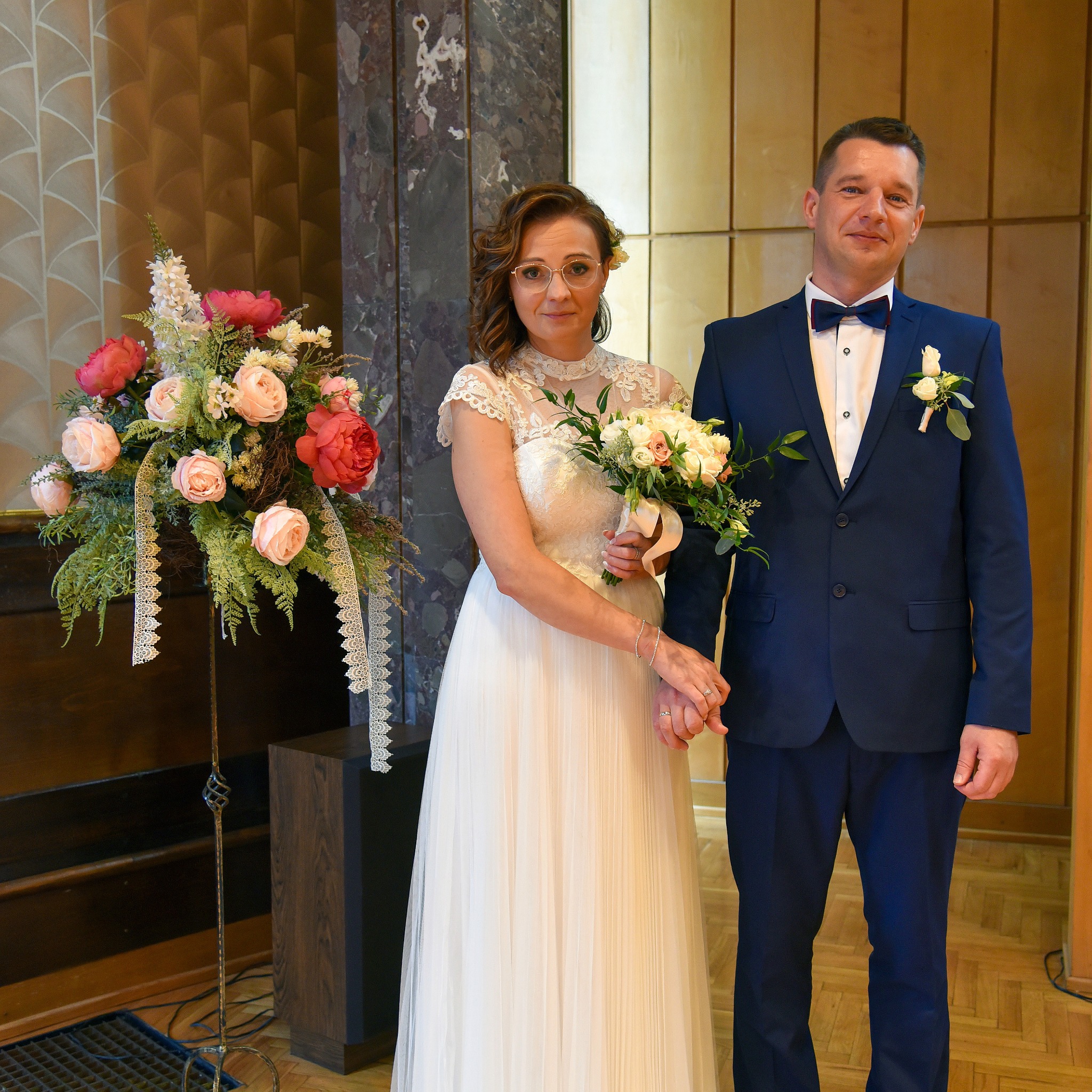 Para młoda w Zawadzie pozuje do zdjęcia w eleganckich strojach ślubnych, trzymając się za ręce, z bukietem kwiatów w dłoni panny młodej i dekoracją kwiatową obok.