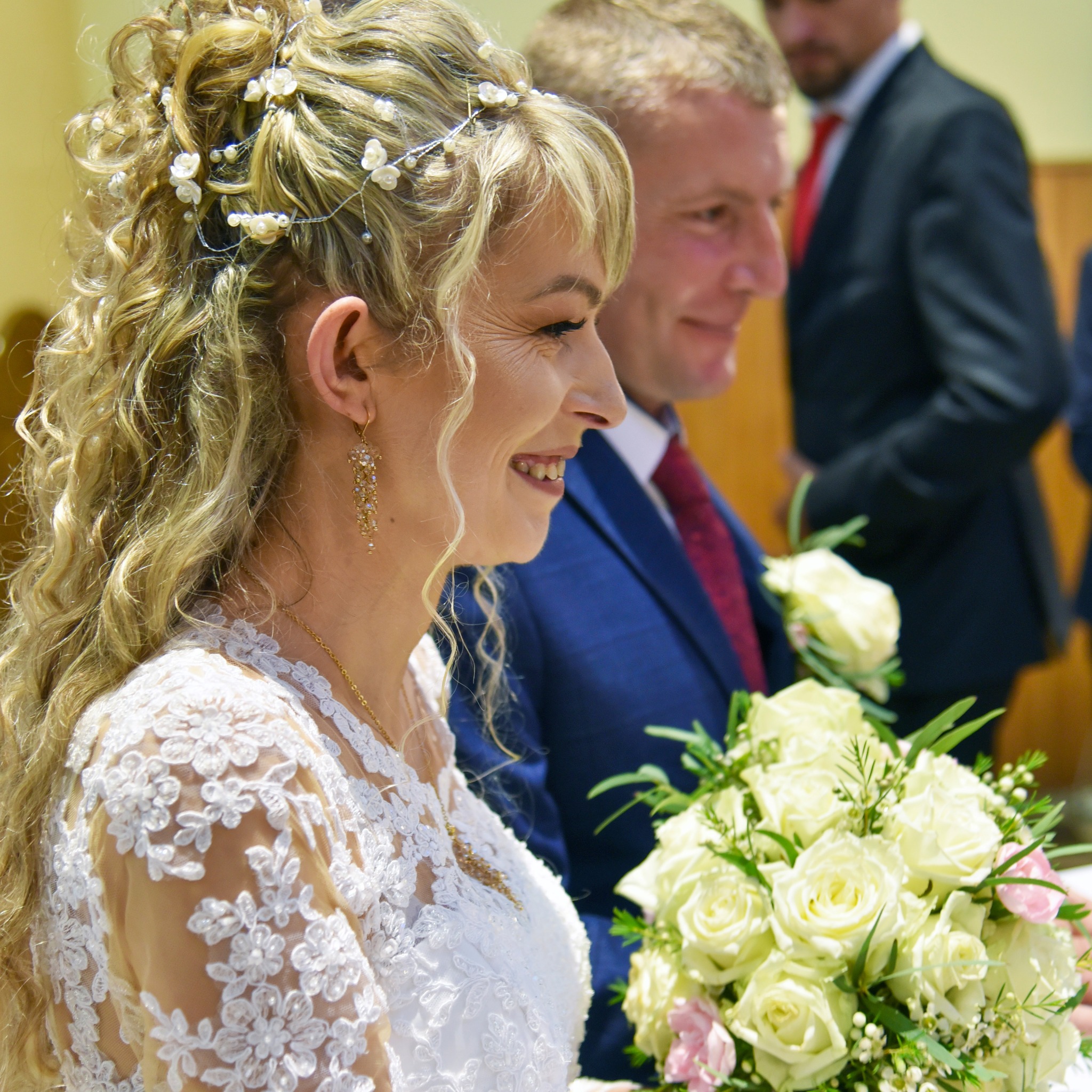 Uśmiechnięta panna młoda w koronkowej sukni z bukietem białych róż, z ozdobami we włosach i złotymi kolczykami, obok pana młodego w granatowym garniturze i czerwonym krawacie.