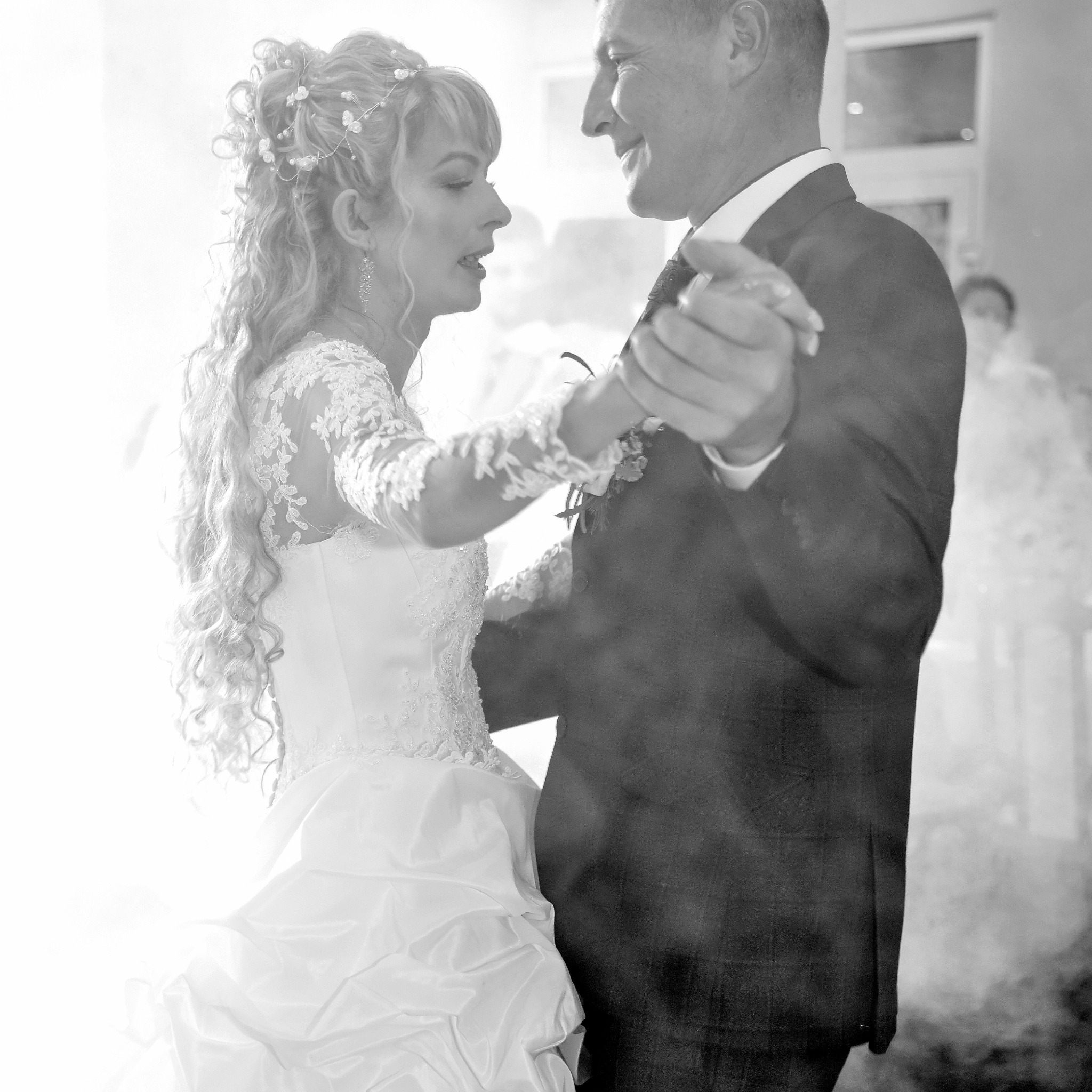 Czarno-białe zdjęcie pary młodej podczas pierwszego tańca, panna młoda w koronkowej sukni z długimi włosami ozdobionymi kwiatami, pan młody w ciemnym garniturze, w tle delikatna mgła.