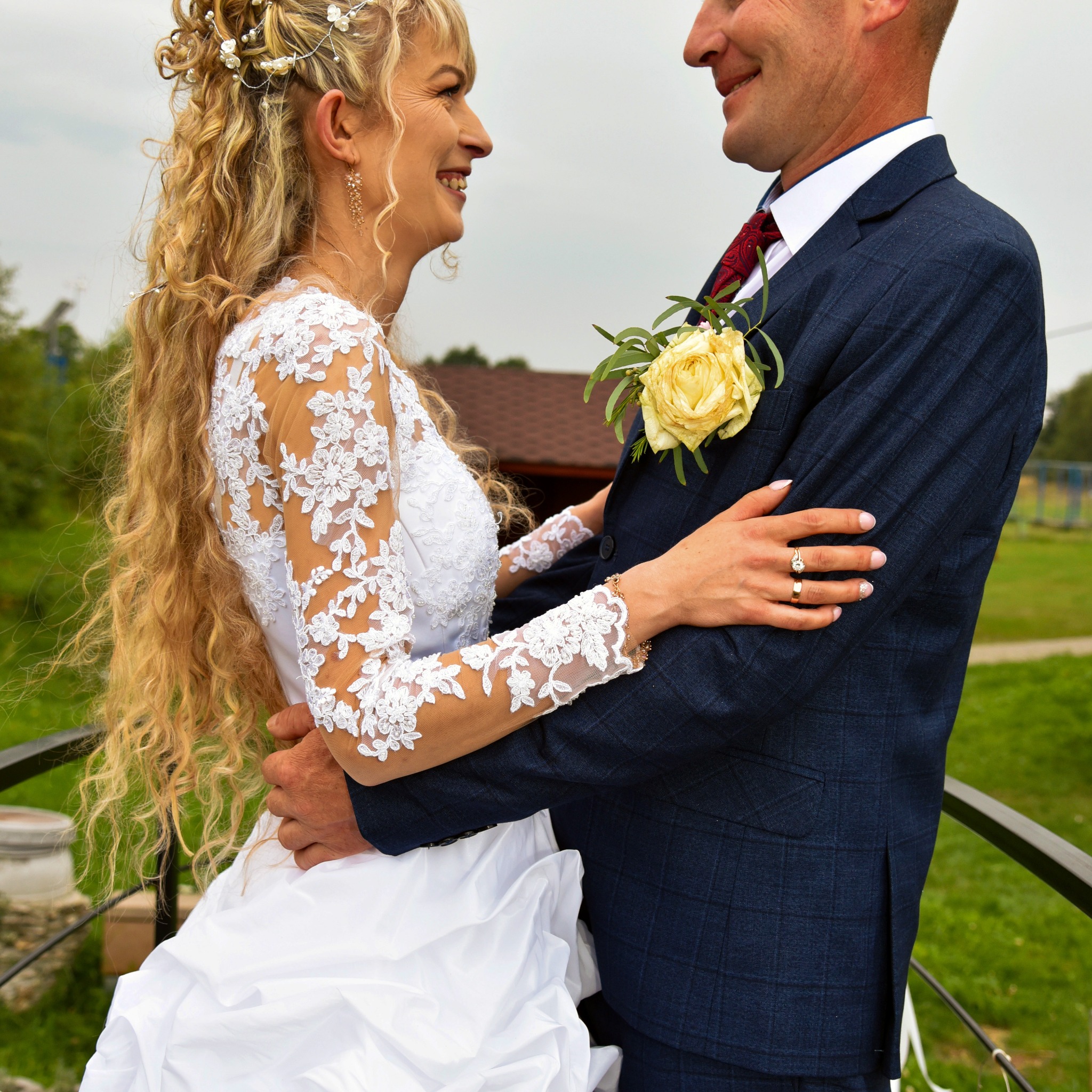 Uśmiechnięta panna młoda w koronkowej sukni spogląda na pana młodego w niebieskim garniturze z żółtą różą w butonierce, widoczne obrączki ślubne.