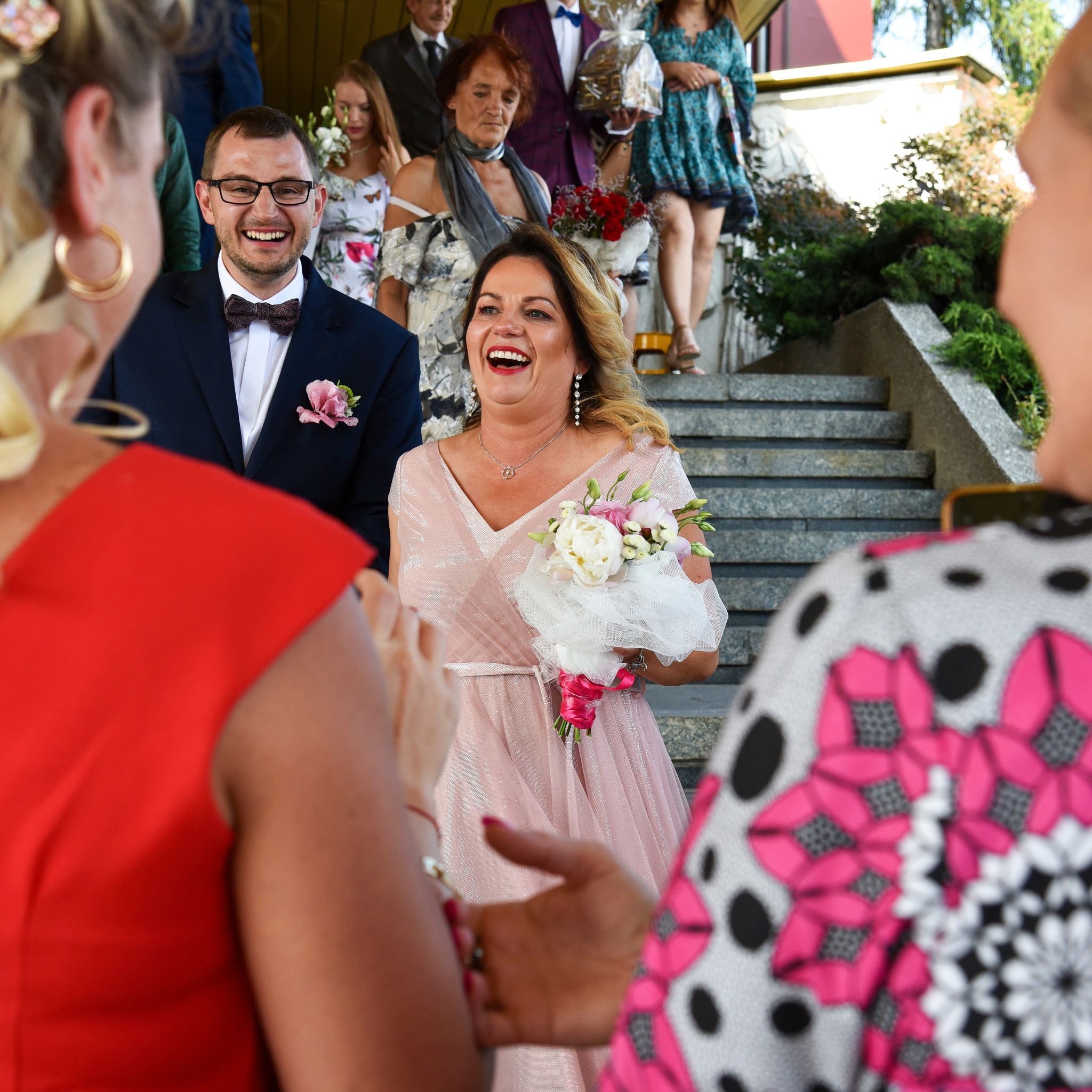 Uśmiechnięta panna młoda w pudrowej sukni z bukietem, schodzi po schodach w otoczeniu gości weselnych.