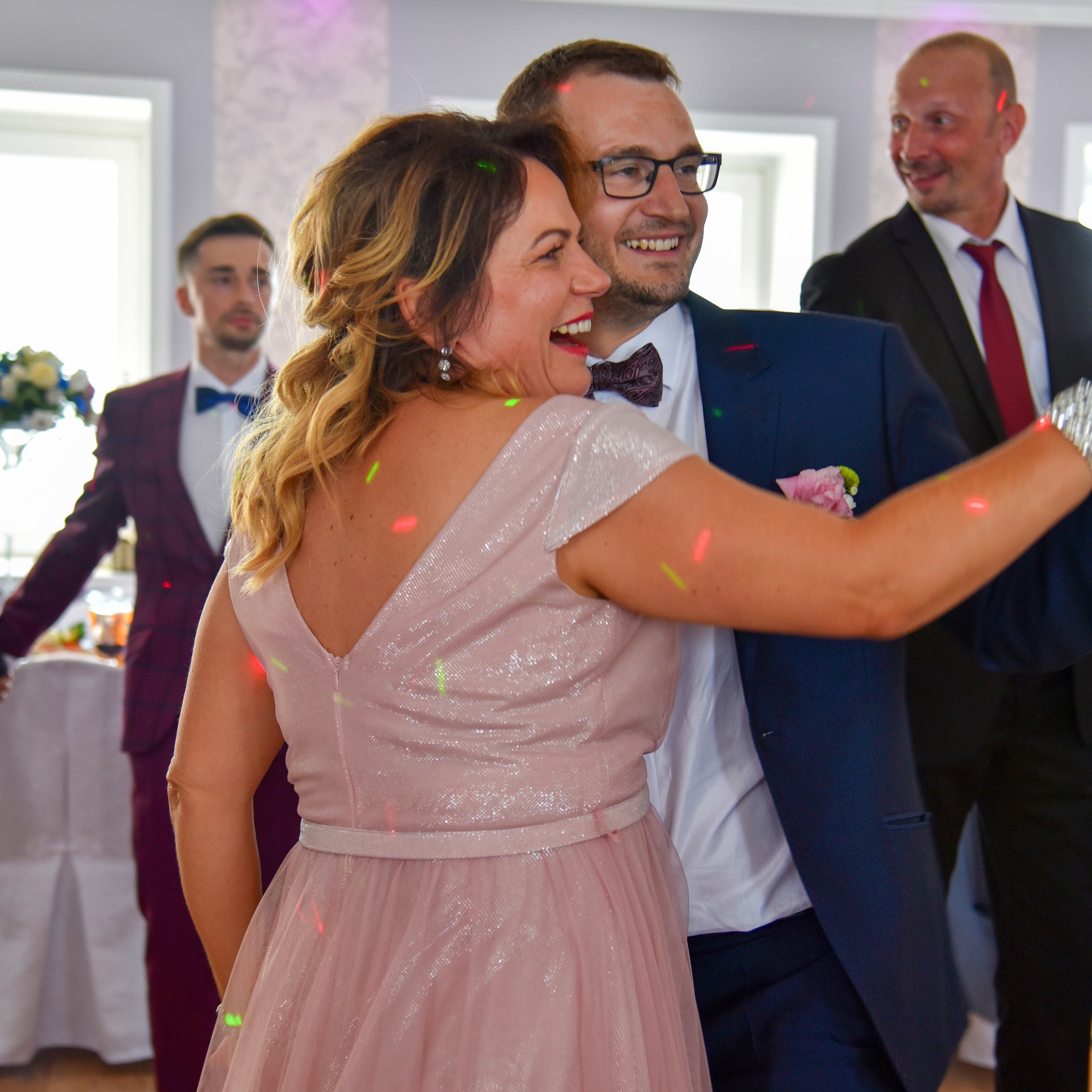 Uśmiechnięta para w strojach wieczorowych tańczy na przyjęciu, w tle widoczni goście w garniturach i dekoracje sali.