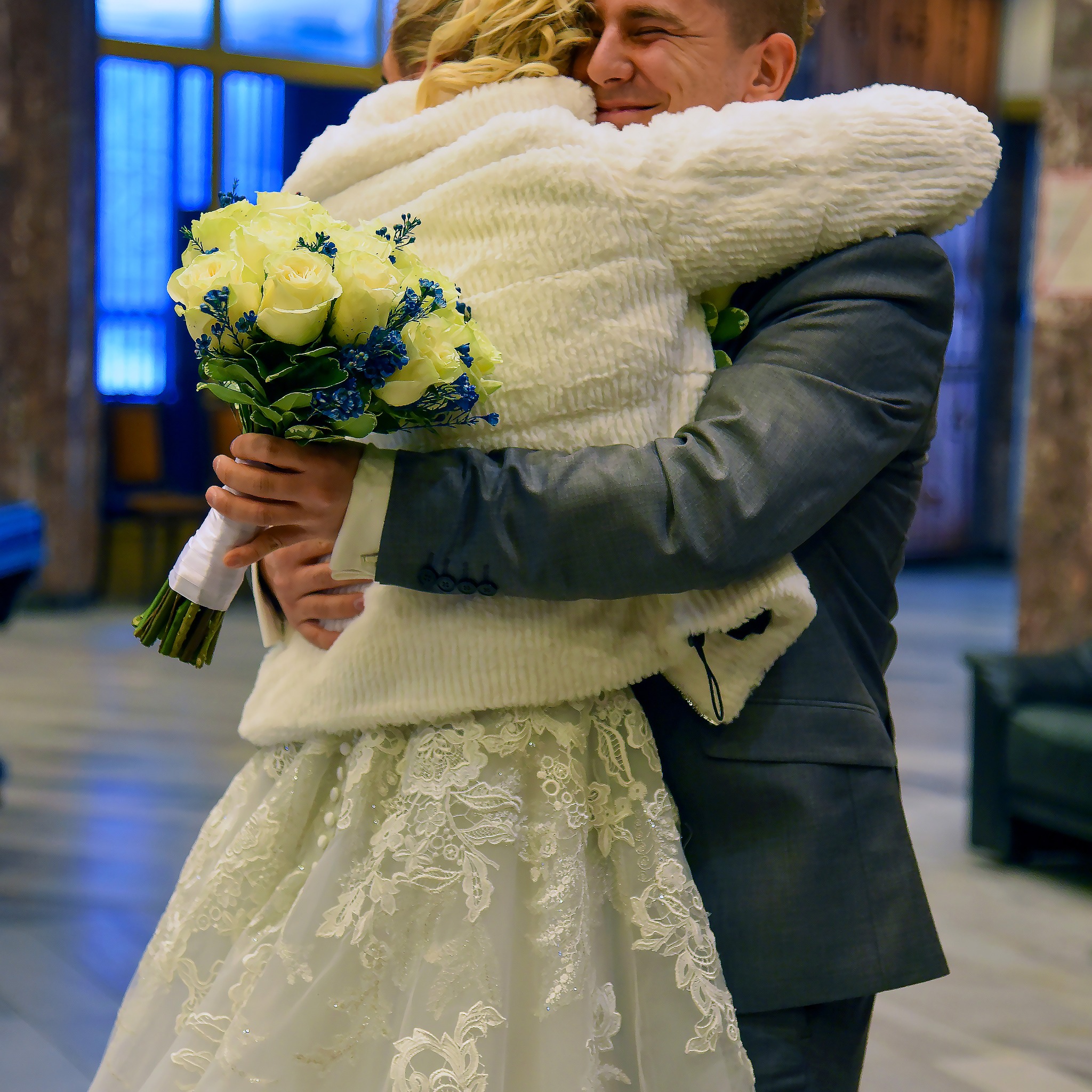 Uśmiechnięty pan młody w szarym garniturze obejmuje pannę młodą w białej sukni z koronkowymi detalami i futrzanym okryciem, trzymającą bukiet białych róż i niebieskich kwiatów.