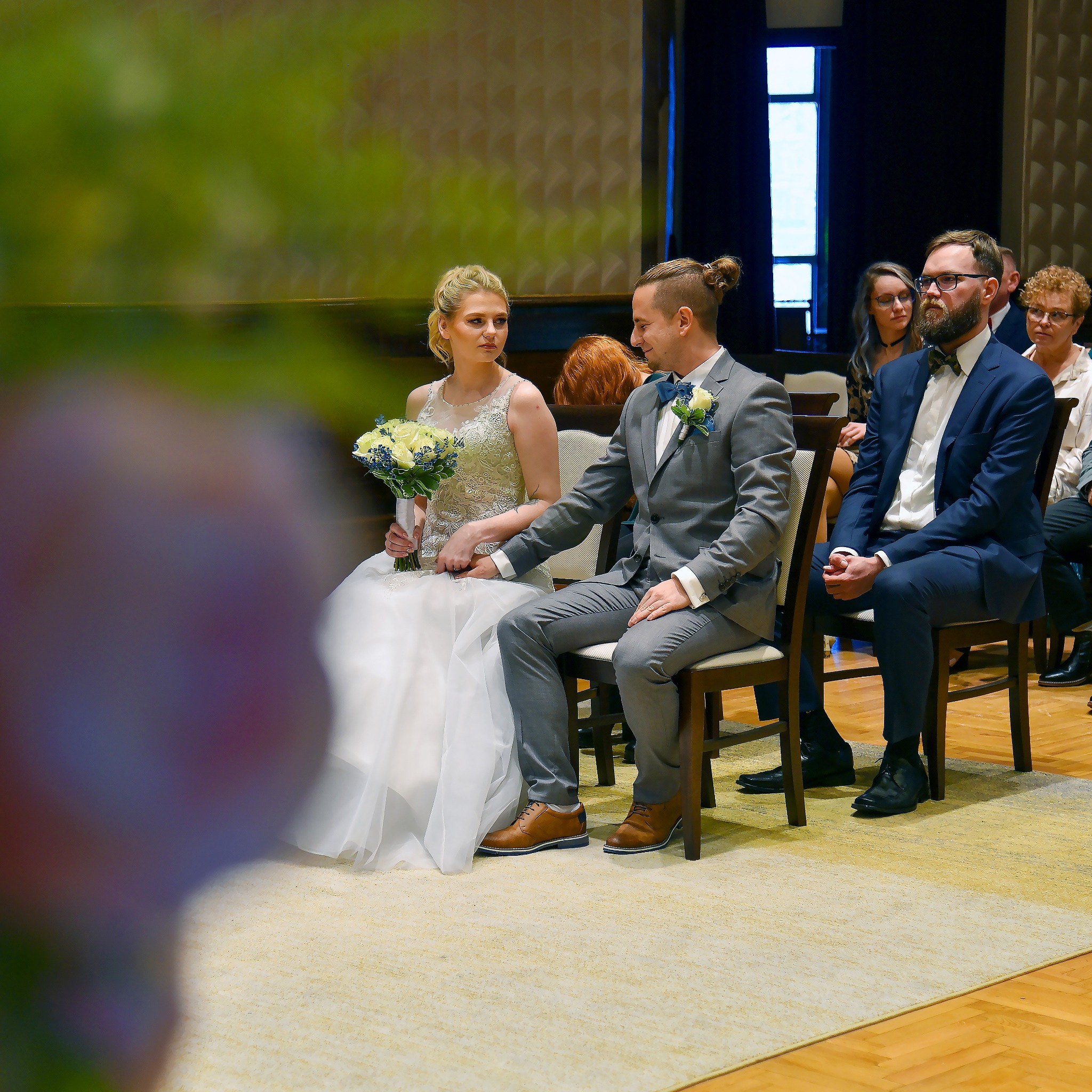 Para młoda siedzi obok siebie podczas ceremonii, panna młoda trzyma bukiet białych róż i niebieskich akcentów, goście w tle.