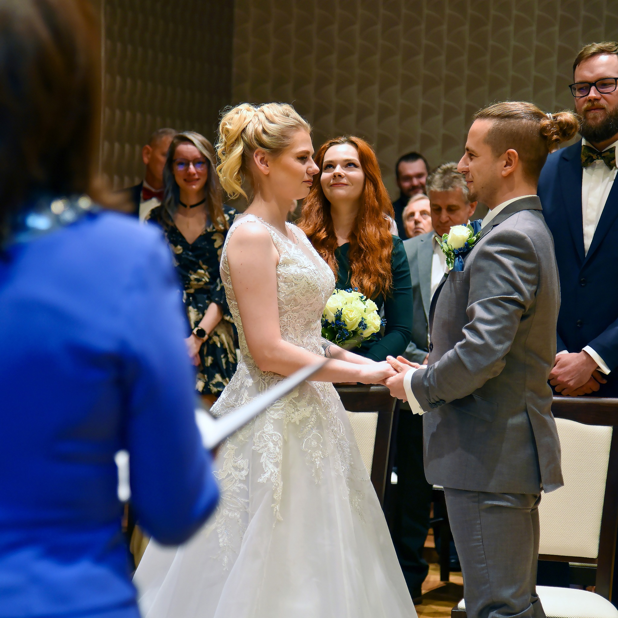 Para młoda trzymająca się za ręce podczas ceremonii ślubnej, panna młoda w koronkowej sukni z bukietem, pan młody w szarym garniturze z butonierką, goście w tle.