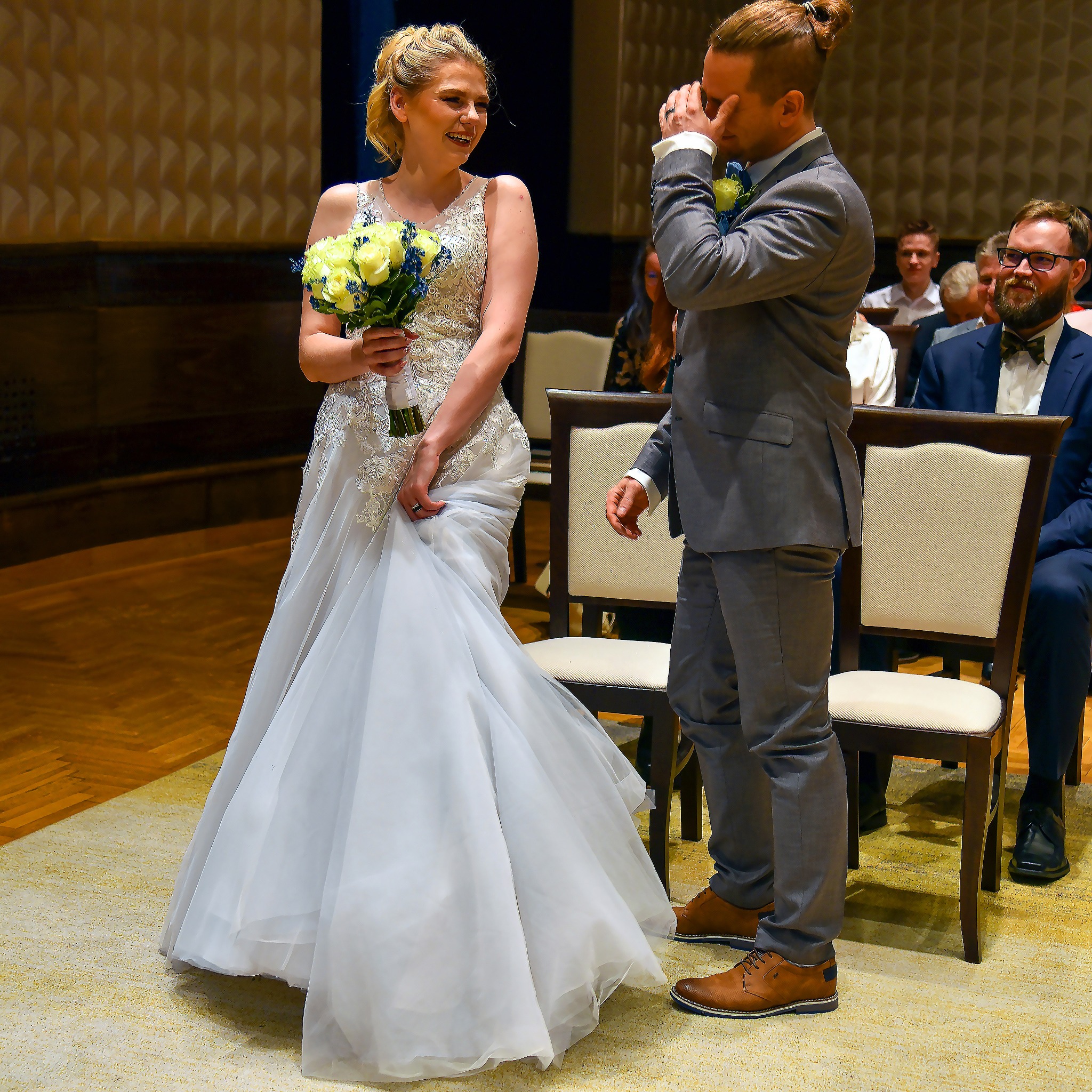 Panna młoda w białej sukni z bukietem żółtych róż uśmiecha się do wzruszonego pana młodego w szarym garniturze podczas ceremonii ślubnej.