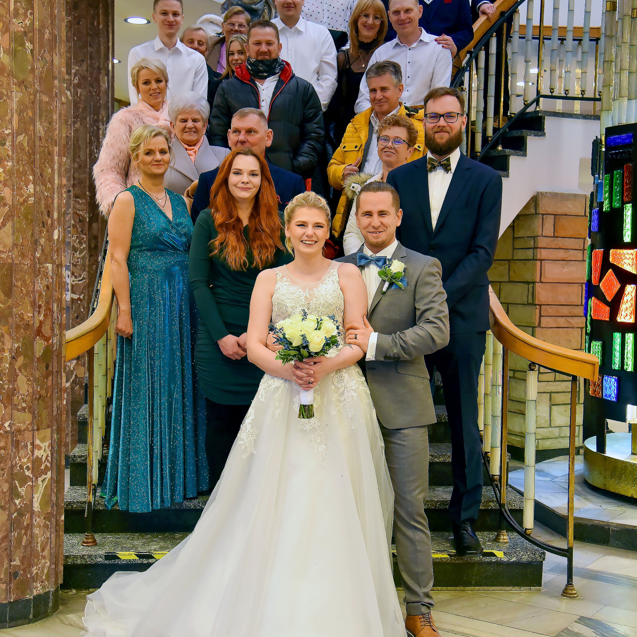 Para młoda pozuje na schodach z gośćmi weselnymi w tle, panna młoda trzyma bukiet białych róż, a pan młody ma szary garnitur i niebieską muszkę.