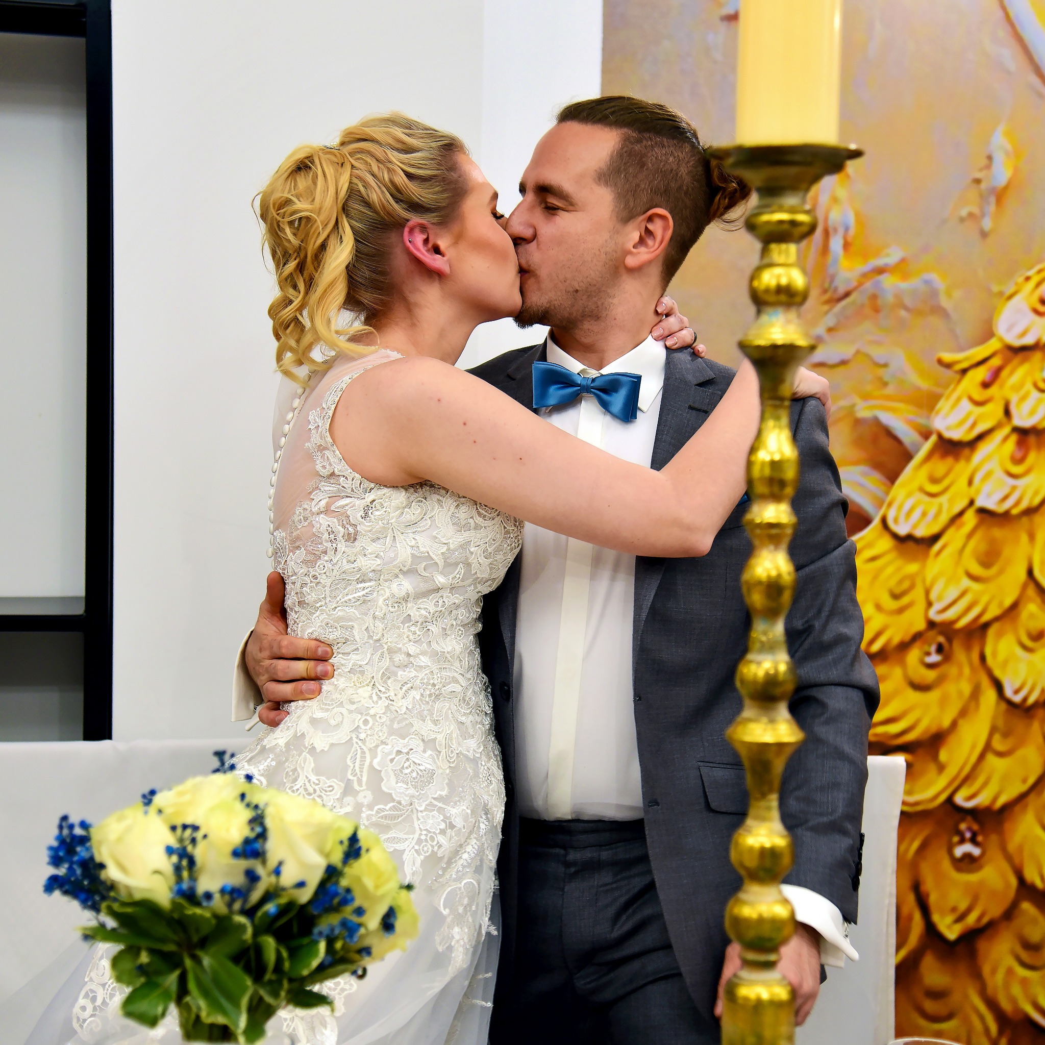 Pocałunek nowożeńców: blondwłosa panna młoda w koronkowej sukni i pan młody w szarym garniturze z błękitną muchą, bukiet białych i niebieskich kwiatów na stole, złoty świecznik i dekoracja za nimi.