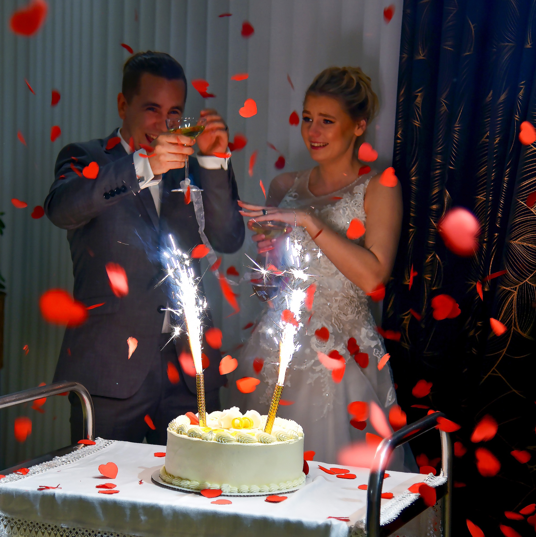 Uśmiechnięta para młoda wznosi toast za tort weselny ze sztucznymi ogniami, otoczona opadającymi konfetti w kształcie serc.
