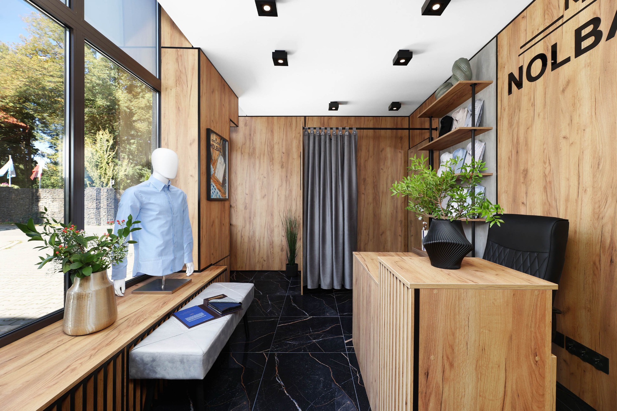 Wnętrze butiku odzieżowego z drewnianymi panelami, manekinem w błękitnej koszuli przy oknie i czarną marmurową podłogą. Na biurku wazon z rośliną.