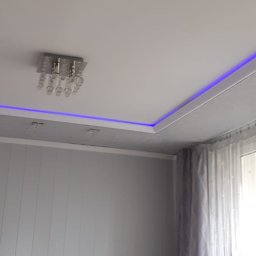 Adam Wykonczenia - Szary salon z podwieszanym sufitem z niebieskim oświetleniem LED i kryształowym żyrandolem, widoczne panele ścienne i zasłony w oknie.