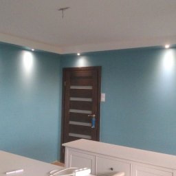 Adam Wykonczenia - Turkusowa ściana z równomiernie rozmieszczonymi punktowymi źródłami światła, widoczna drewniana ościeżnica drzwi z szybami i fragment białych mebli w dolnej części kadru.