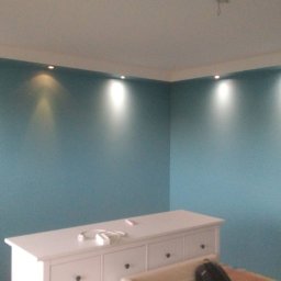 Adam Wykonczenia - Ściana pomalowana na niebiesko z oświetleniem punktowym w suficie, biała komoda z gniazdkiem i ładowarką na górze, widok na róg pokoju.
