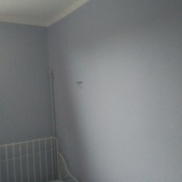 Adam Wykonczenia - Świeżo pomalowana ściana na szaro w pokoju z widocznym fragmentem okna, zasłony w czerwone wzory i metalowego zagłówka łóżka.