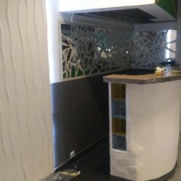 Adam Wykonczenia - Nowoczesna ściana z trójwymiarową fakturą obok baru z mozaikową górą i kolorowymi wstawkami, widok na okno i dekoracyjną ściankę ażurową w tle.