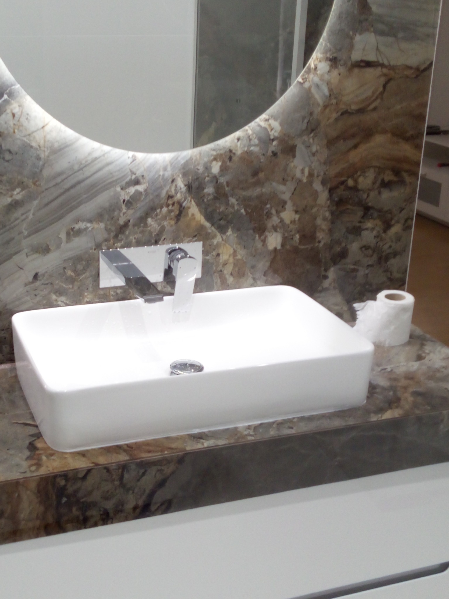 Biała, prostokątna umywalka nablatowa z chromowaną baterią i odpływem, zamontowana na blacie z marmurowym wzorem, obok rolka papieru toaletowego, widoczne okrągłe lustro z podświetleniem.