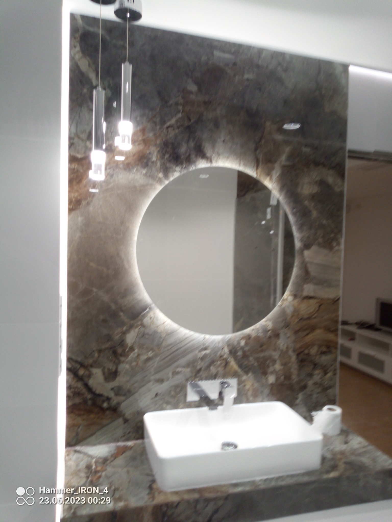 Nowoczesna łazienka z umywalką nablatową, okrągłym lustrem z podświetleniem LED i marmurową ścianą w odcieniach szarości i brązu, uzupełniona minimalistycznymi lampami wiszącymi.