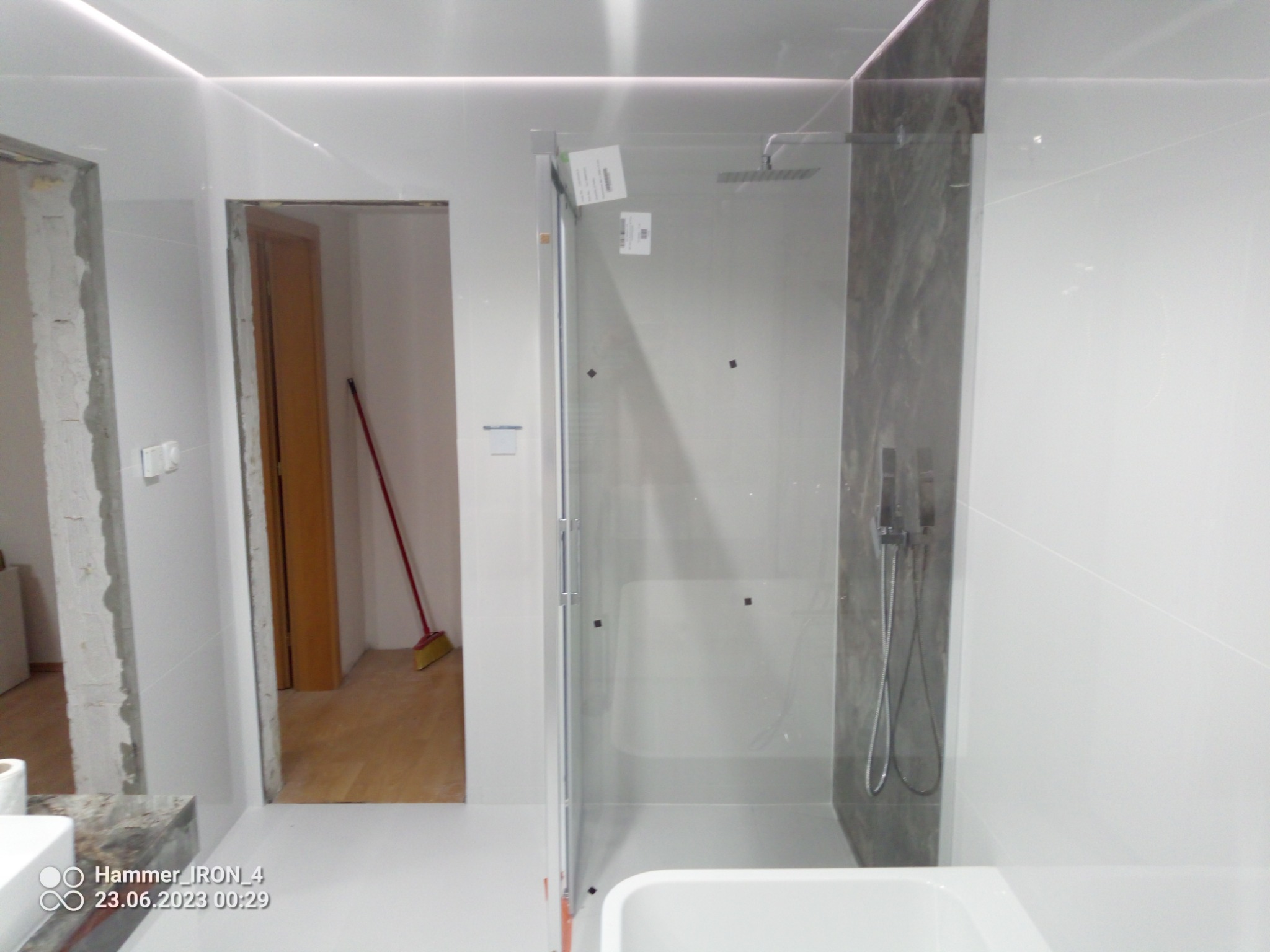 Nowoczesna kabina prysznicowa z szarym panelem imitującym marmur, widoczna podczas montażu w łazience z białymi ścianami i podświetleniem LED, obok niedokończone futryny drzwi i widoczna miotła...