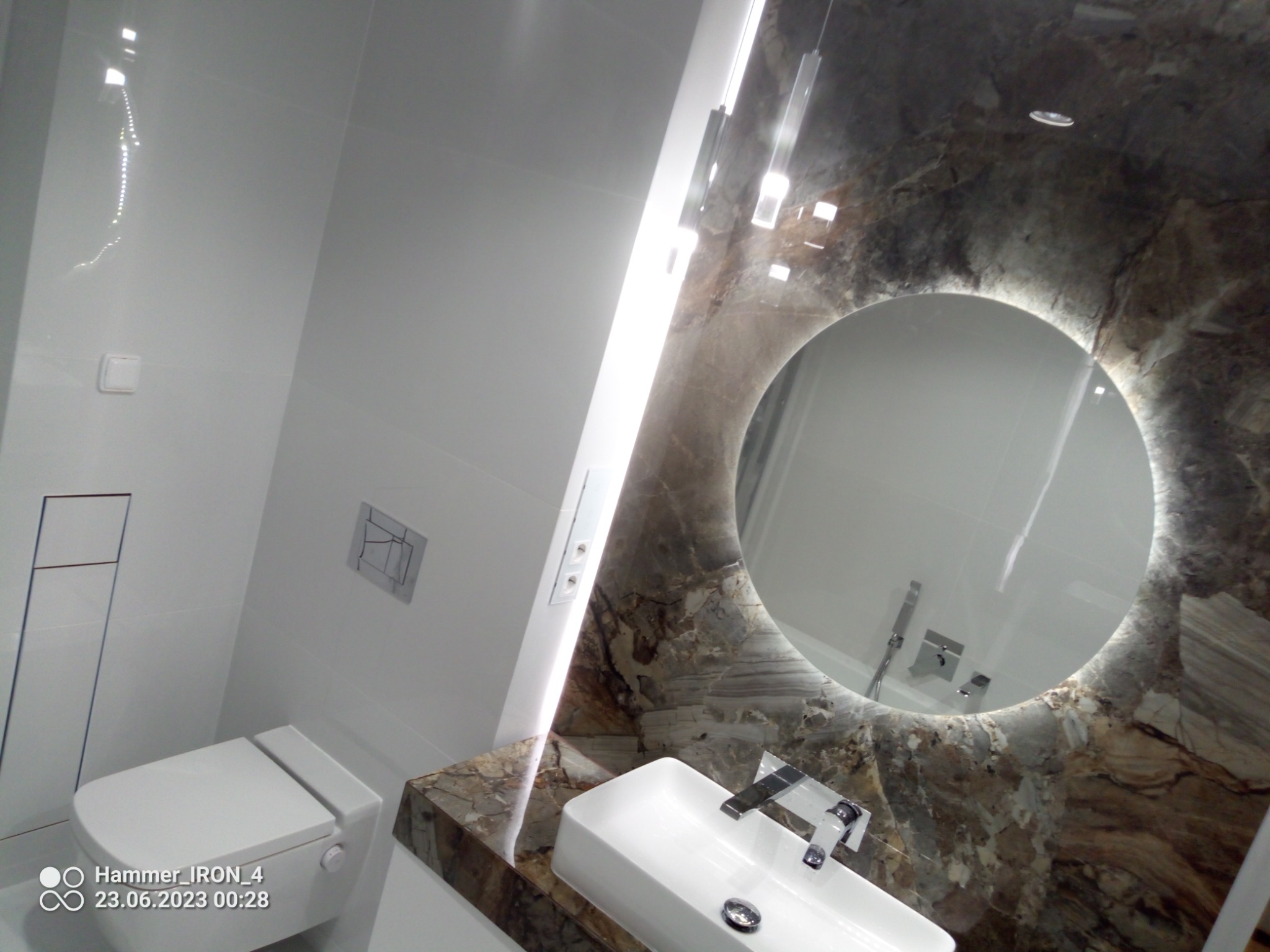 Nowoczesna łazienka z marmurowym blatem i okrągłym lustrem z podświetleniem LED, biała ceramika sanitarna i minimalistyczna armatura.