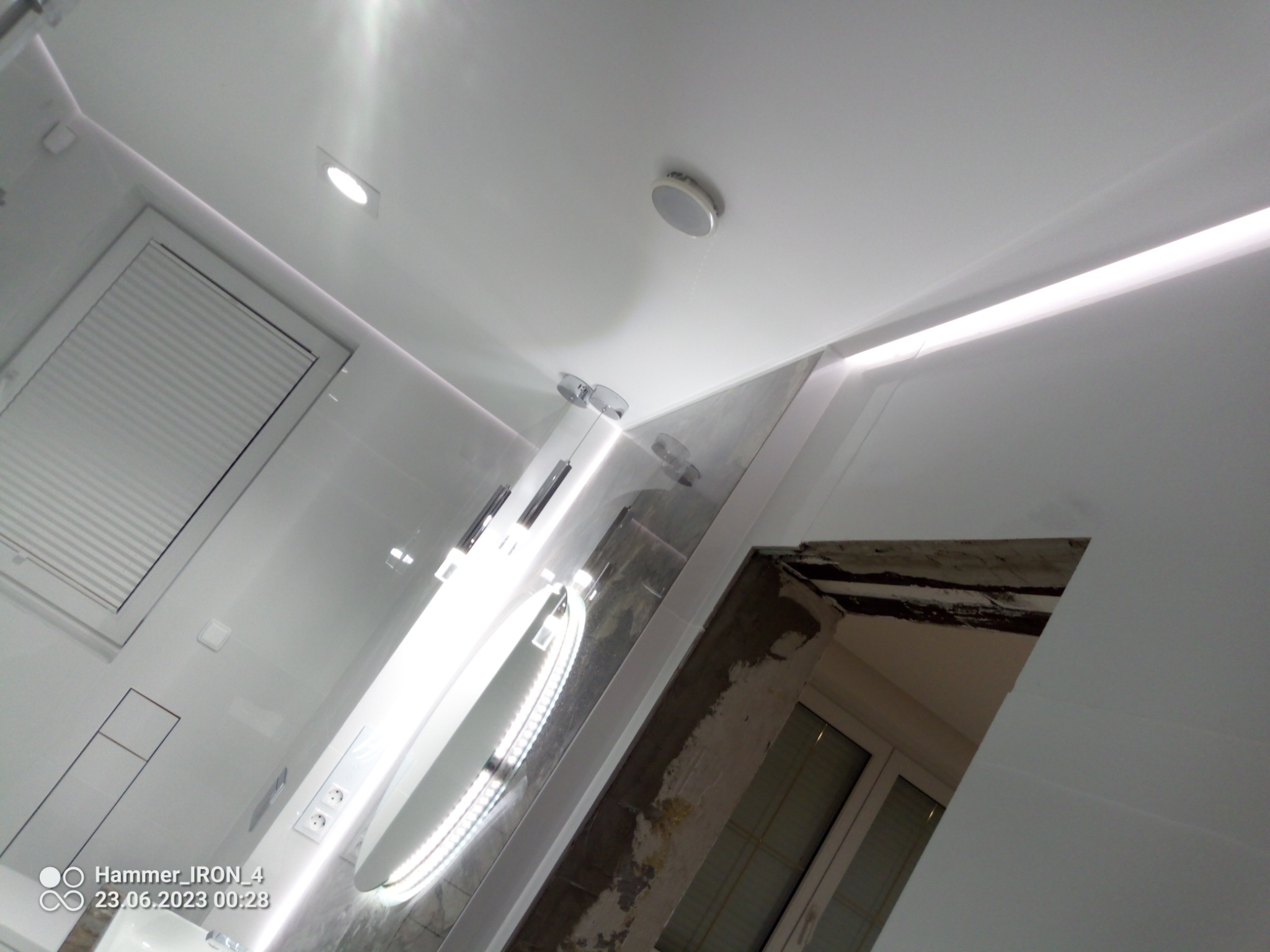 Wnętrze łazienki w trakcie remontu, widoczne białe płytki, okrągłe lustro z podświetleniem LED, sufit z oświetleniem punktowym i ledowym, fragment okna oraz ściana w stanie surowym przed wykończeniem.