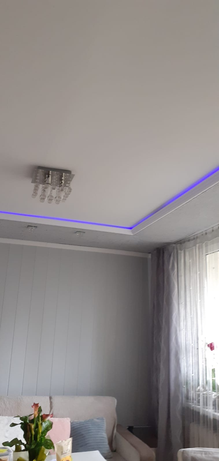 Szary salon z podwieszanym sufitem z niebieskim oświetleniem LED i kryształowym żyrandolem, widoczne panele ścienne i zasłony w oknie.