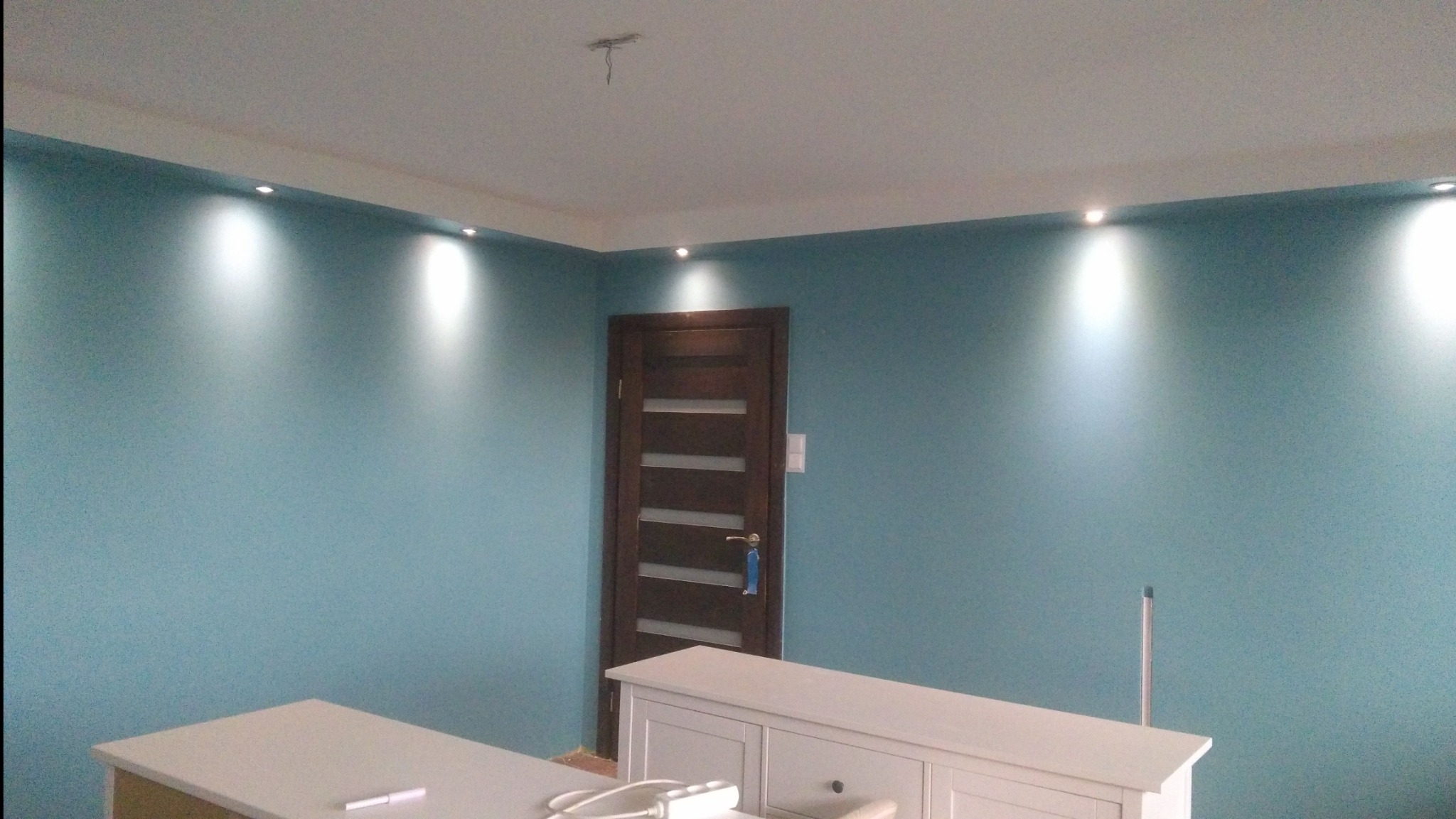 Turkusowa ściana z równomiernie rozmieszczonymi punktowymi źródłami światła, widoczna drewniana ościeżnica drzwi z szybami i fragment białych mebli w dolnej części kadru.