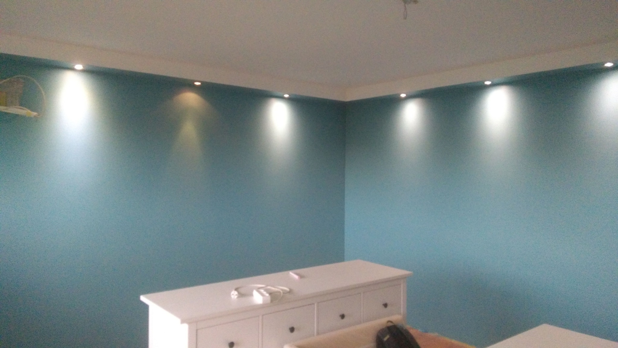 Ściana pomalowana na niebiesko z oświetleniem punktowym w suficie, biała komoda z gniazdkiem i ładowarką na górze, widok na róg pokoju.